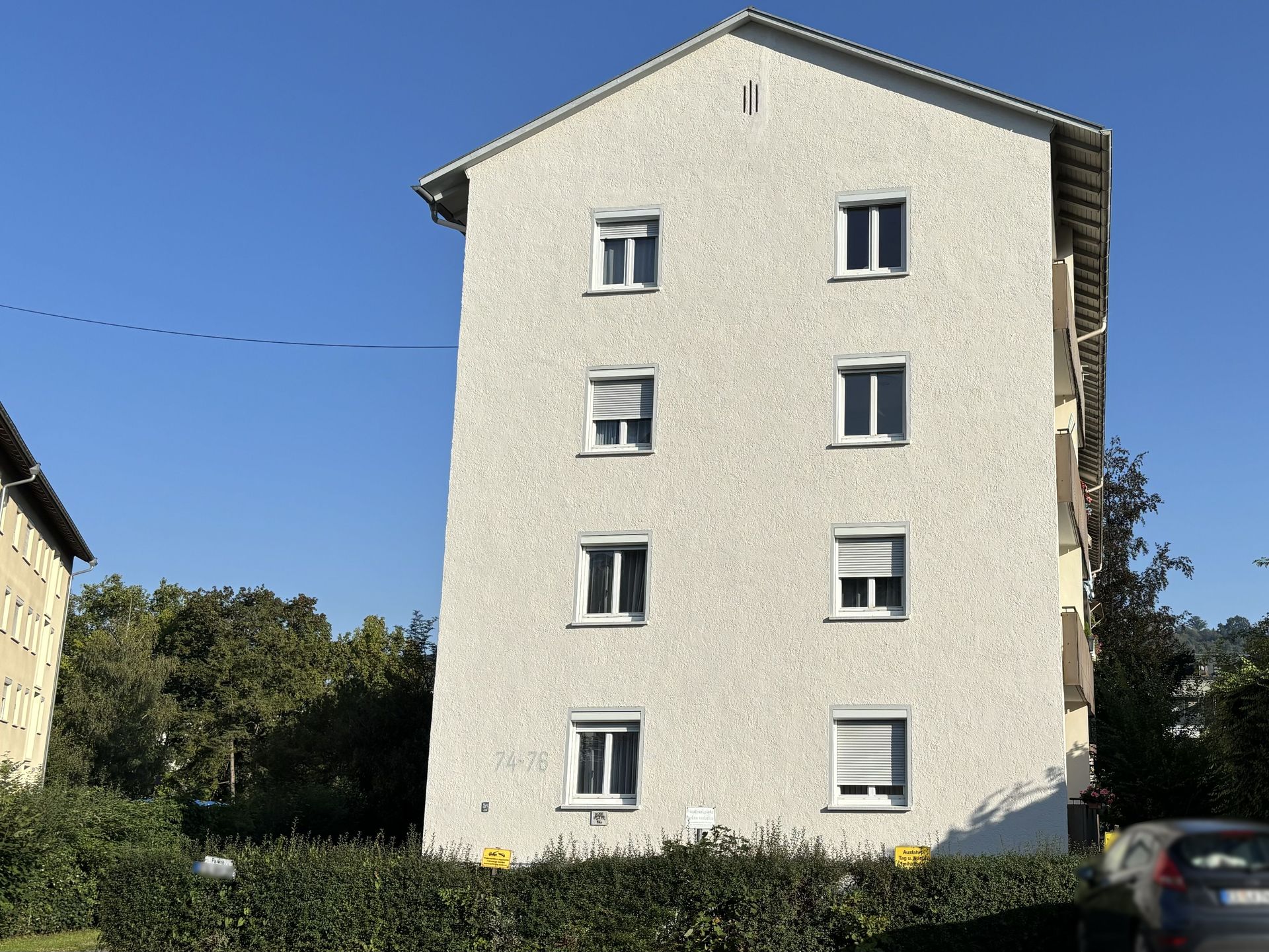 Das Bild zeigt ein Mehrfamilienhaus mit heller Fassade. Vor dem Haus befindet sich eine grüne Hecke und am Straßenrand parkt ein Auto.