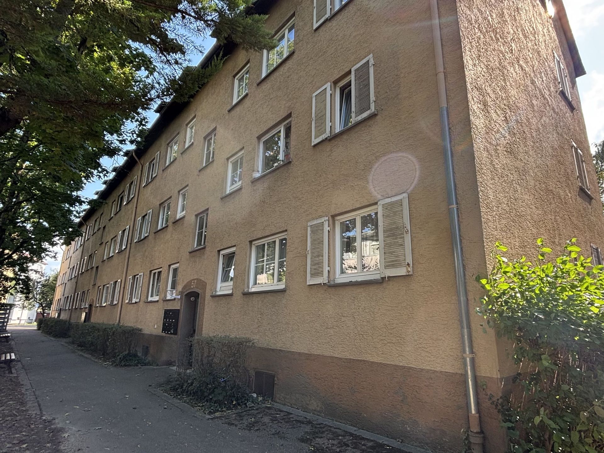 Das Bild zeigt ein Mehrfamilienhaus mit heller Fassade und weißen Fensterläden, das an einem von Bäumen gesäumten Weg liegt.