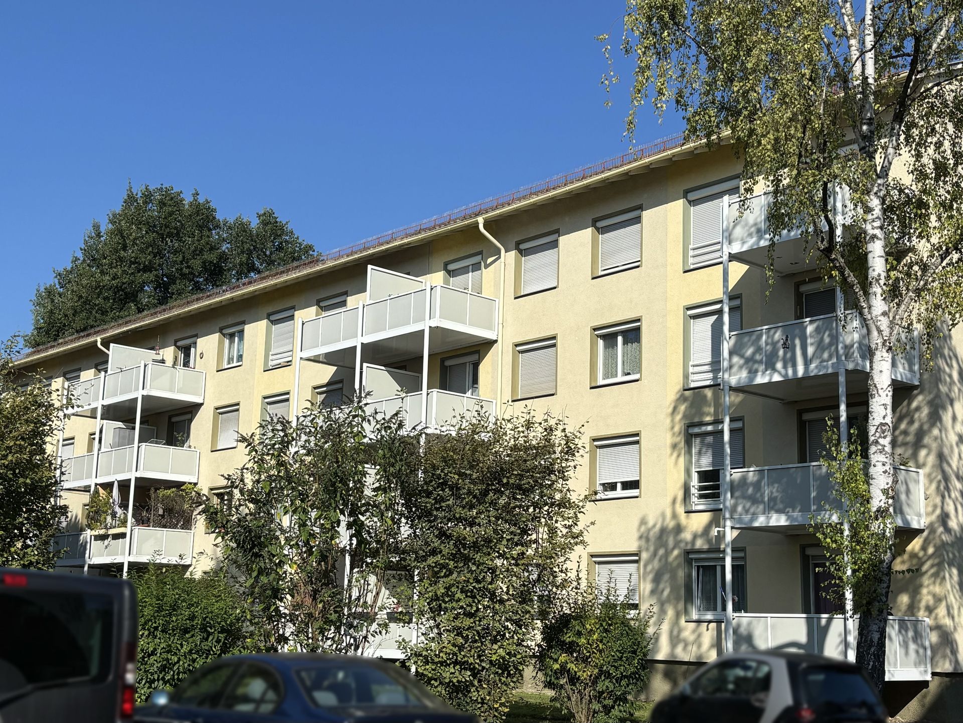 Das Bild zeigt ein Mehrfamilienhaus mit heller Fassade und weißen Balkonen. Vor dem Haus stehen einige Bäume, und am Straßenrand parken Autos.