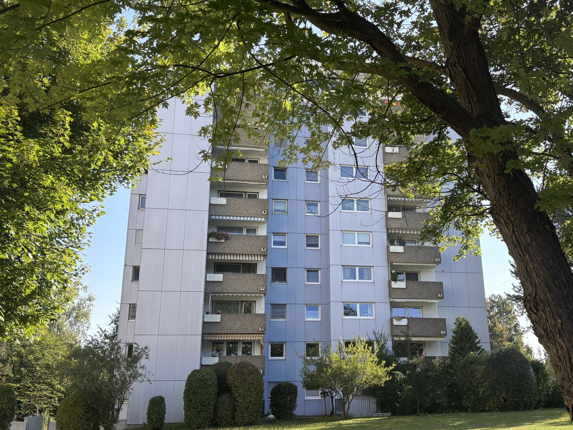 Das Bild zeigt ein hohes Wohngebäude mit abwechselnd hellblauen und weißen Fassadenelementen und vielen Balkonen. Das Gebäude ist von Grün umgeben, mit Bäumen im Vordergrund, die das Haus teilweise verdecken, und Sträuchern am Fuße des Gebäudes.