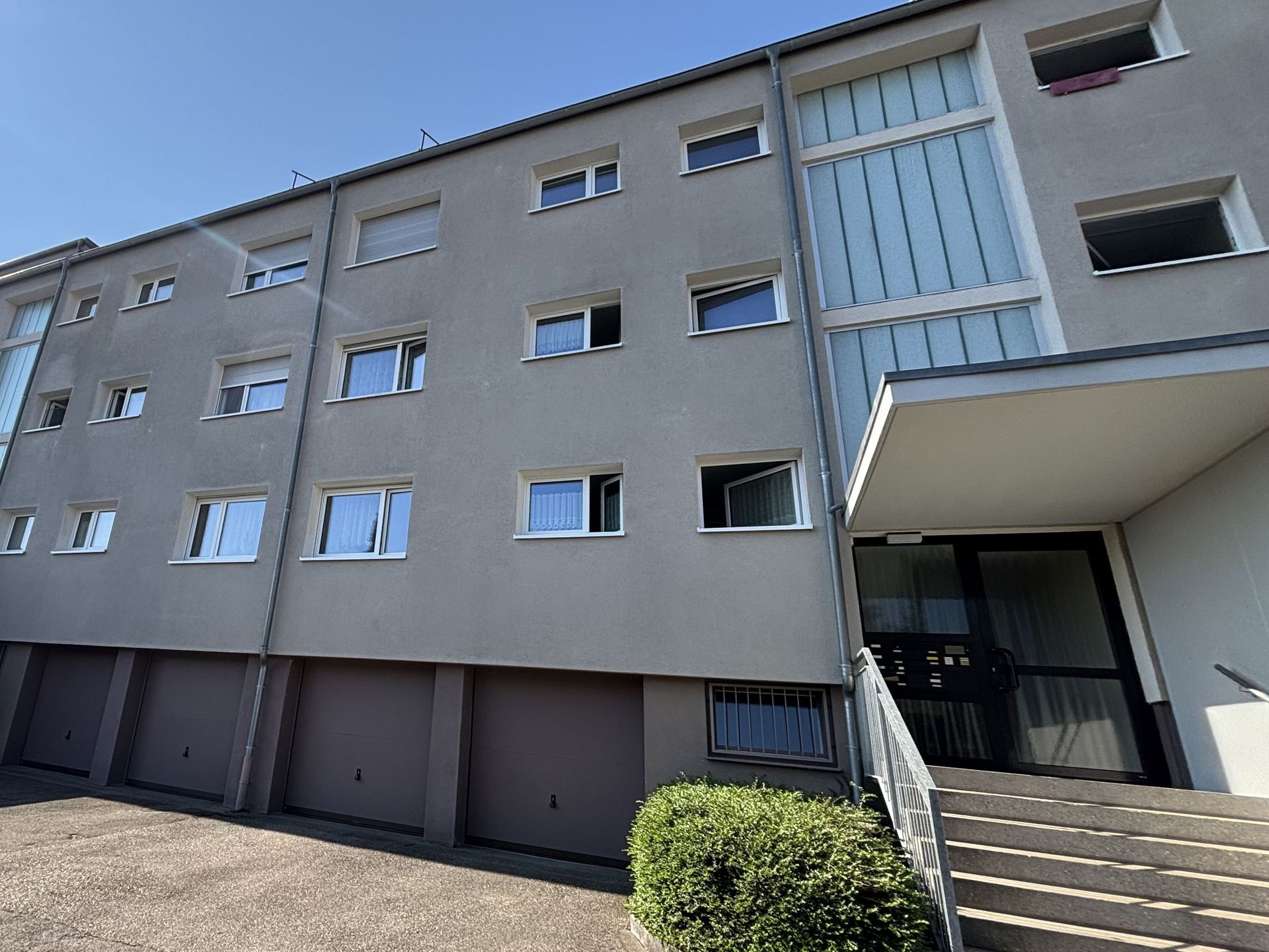 Das Bild zeigt ein Mehrfamilienhaus mit grauer Fassade. Im Erdgeschoss befinden sich Garagen und eine Treppe führt zum Hauseingang.