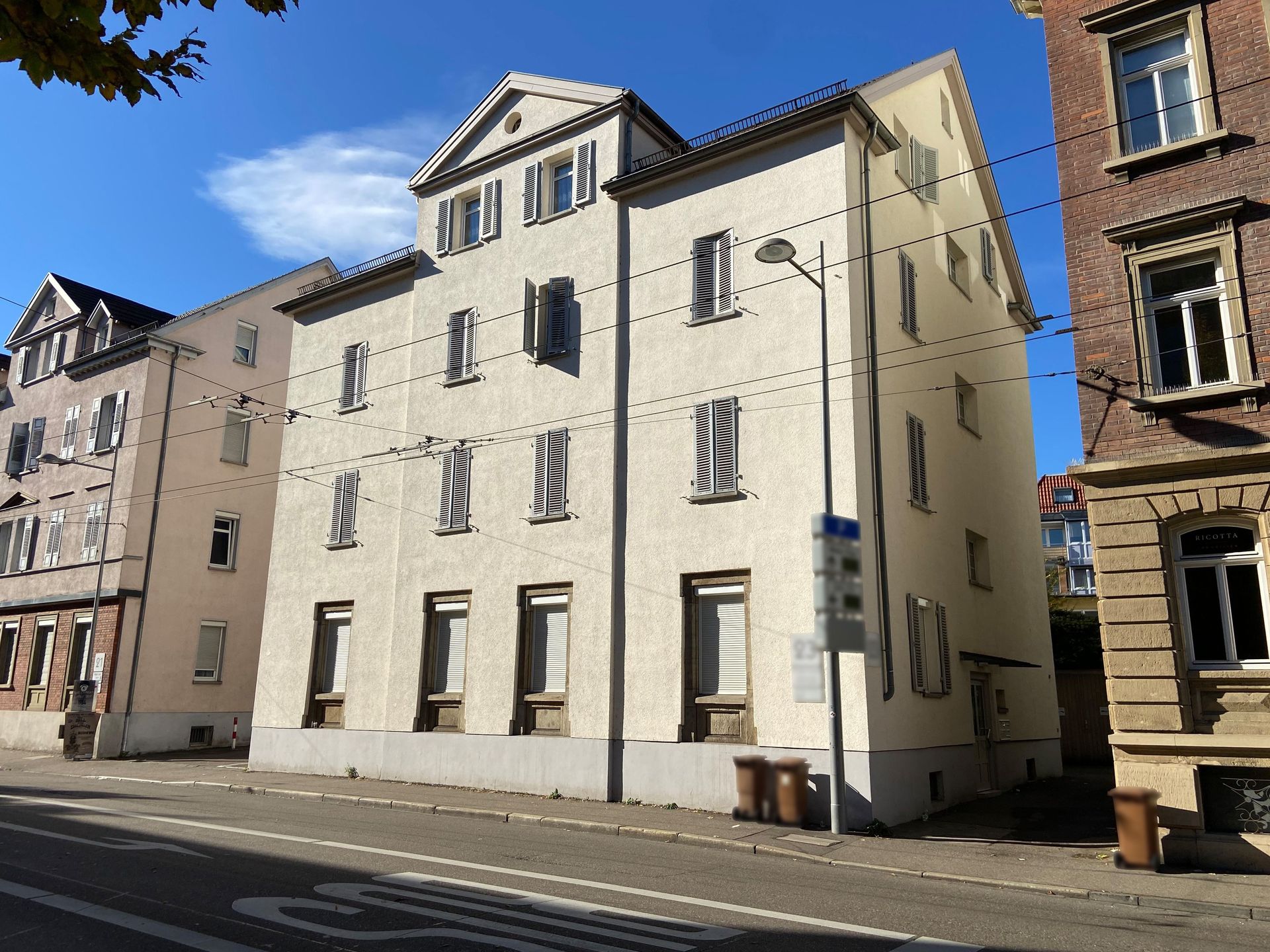 Das Bild zeigt ein Mehrfamilienhaus mit heller Fassade, das an einer Straße mit Oberleitungen steht. Das Gebäude hat mehrere Stockwerke mit zahlreichen Fenstern.