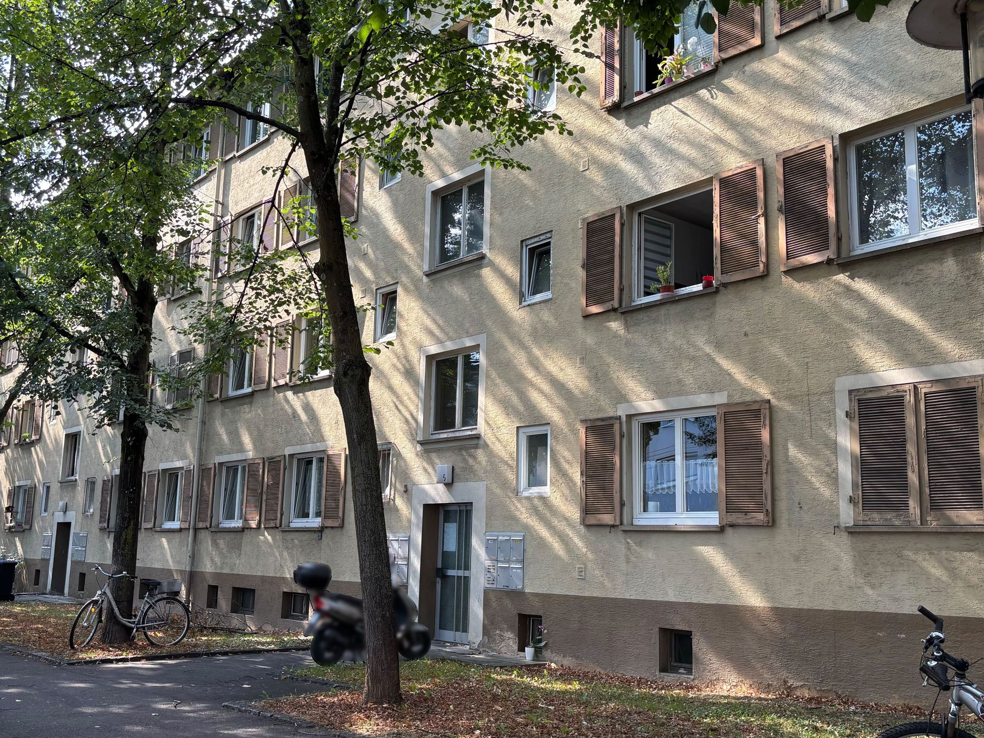 Das Bild zeigt ein Mehrfamilienhaus mit heller Fassade und braunen Fensterläden, vor dem Fahrräder und ein Motorroller geparkt sind. Das Gebäude wird teilweise von Bäumen verdeckt, die Schatten auf die Fassade werfen.