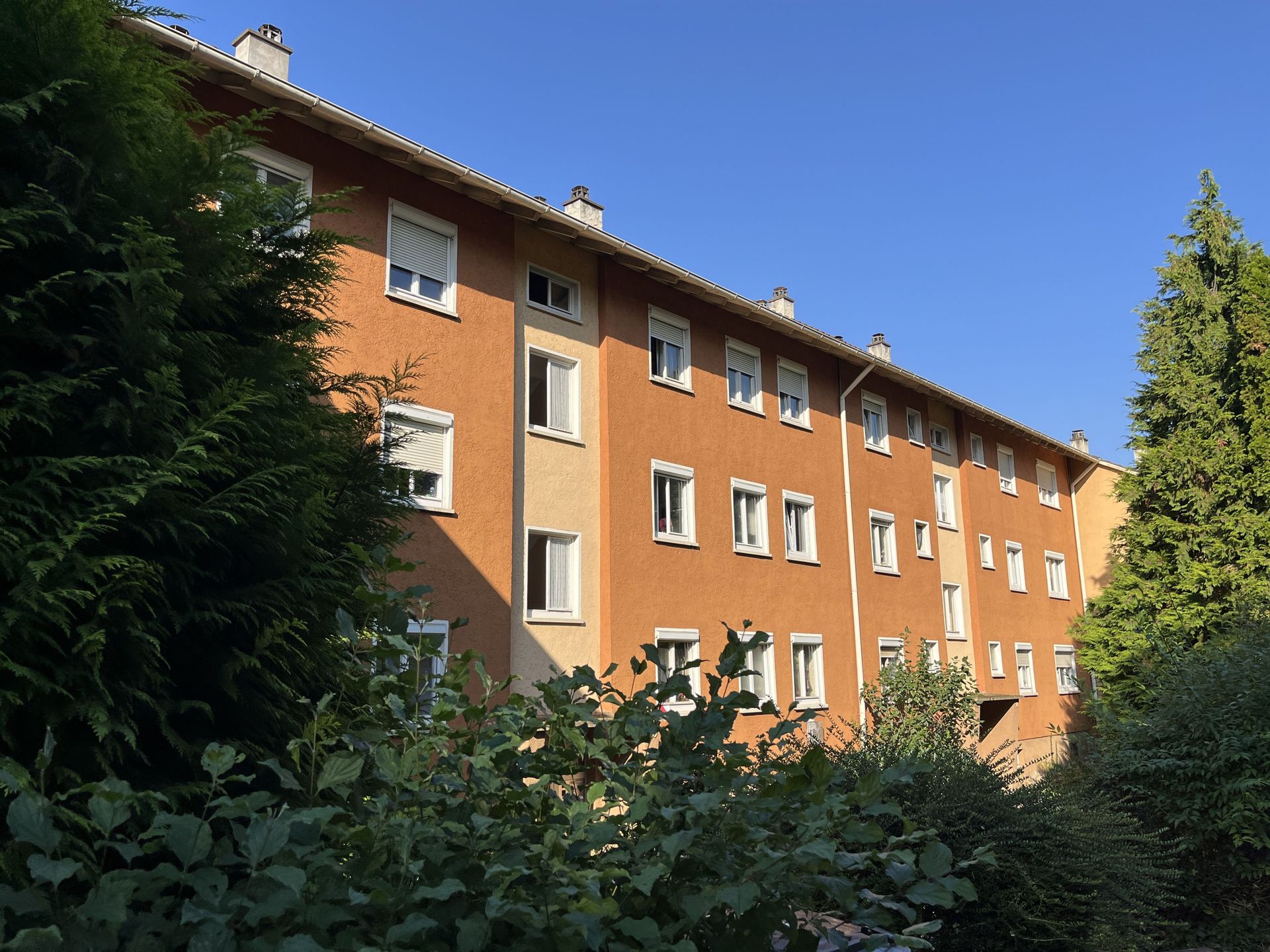 Das Bild zeigt ein Mehrfamilienhaus mit hellbrauner Fassade und weißen Fensterrahmen.  Das Gebäude ist von Bäumen und Sträuchern umgeben, die ihm einen grünen Rahmen verleihen.