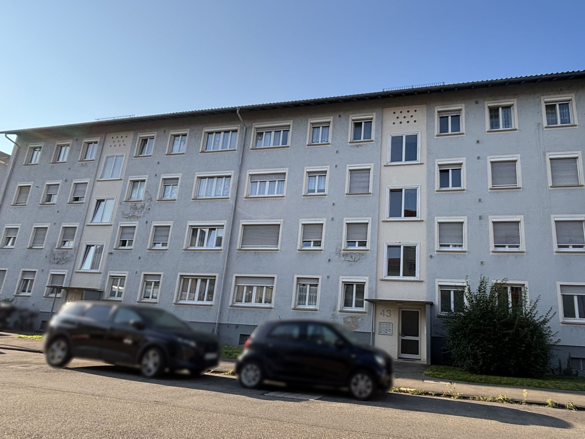 Das Bild zeigt ein Mehrfamilienhaus mit hellblauer Fassade und mehreren Stockwerken. Vor dem Haus stehen zwei Autos am Straßenrand.