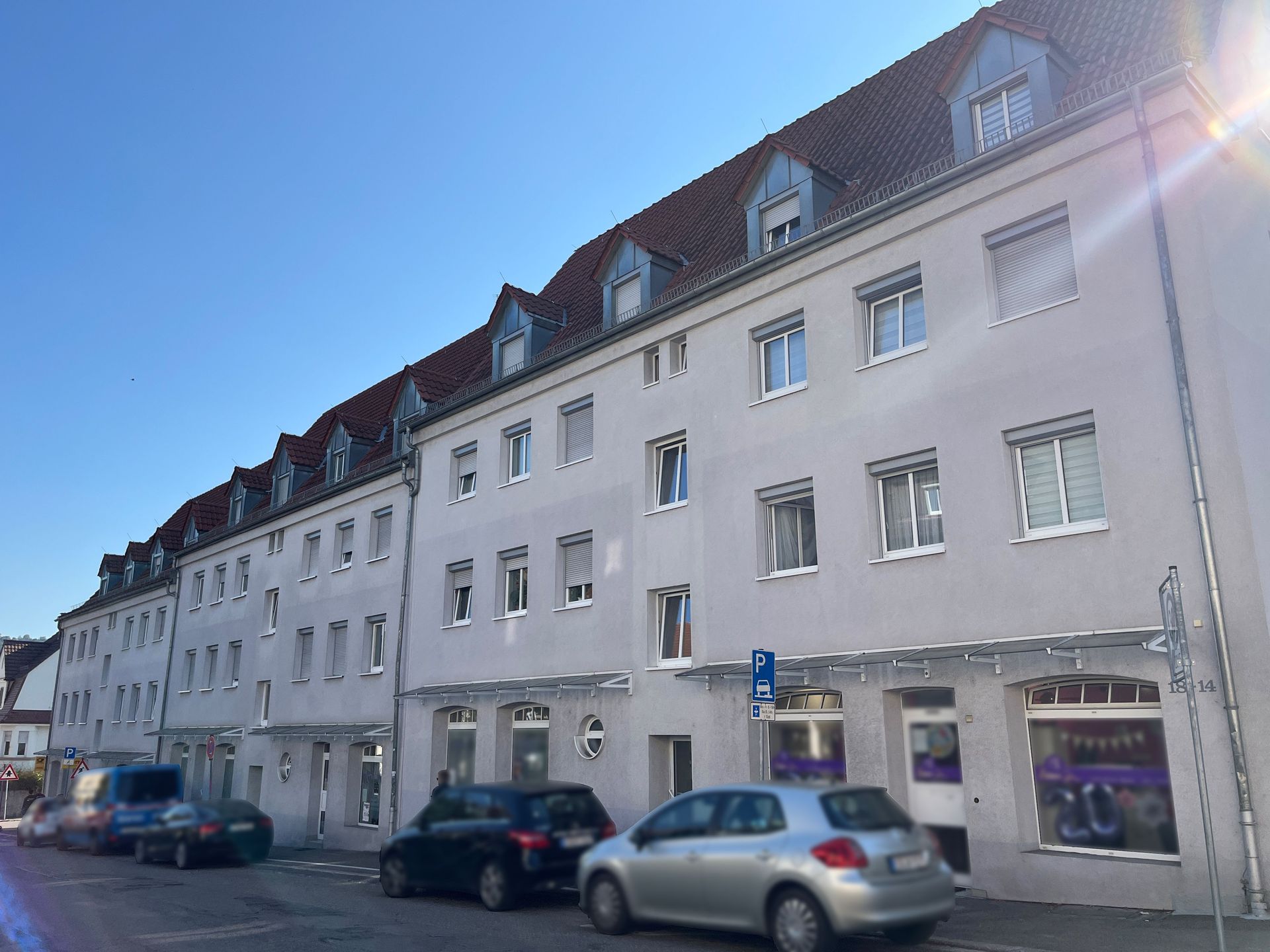 Das Bild zeigt ein Mehrfamilienhaus mit heller Fassade, rotem Ziegeldach und mehreren Dachgauben. Im Erdgeschoss befinden sich Ladenlokale, davor parken Autos am Straßenrand.