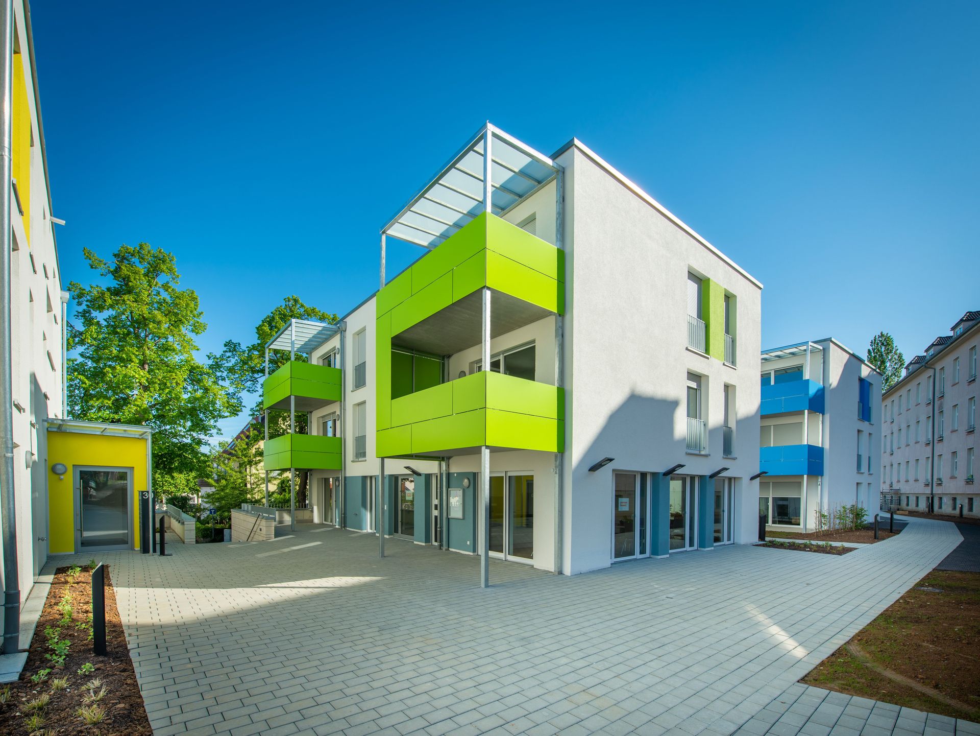 Das Bild zeigt eine Reihe von modernen Reihenhäusern mit weißen Fassaden und Balkonen in leuchtenden Farben, wie grün und blau. Die Häuser gruppieren sich um einen gepflasterten Innenhof und sind von Grünflächen und Bäumen umgeben.