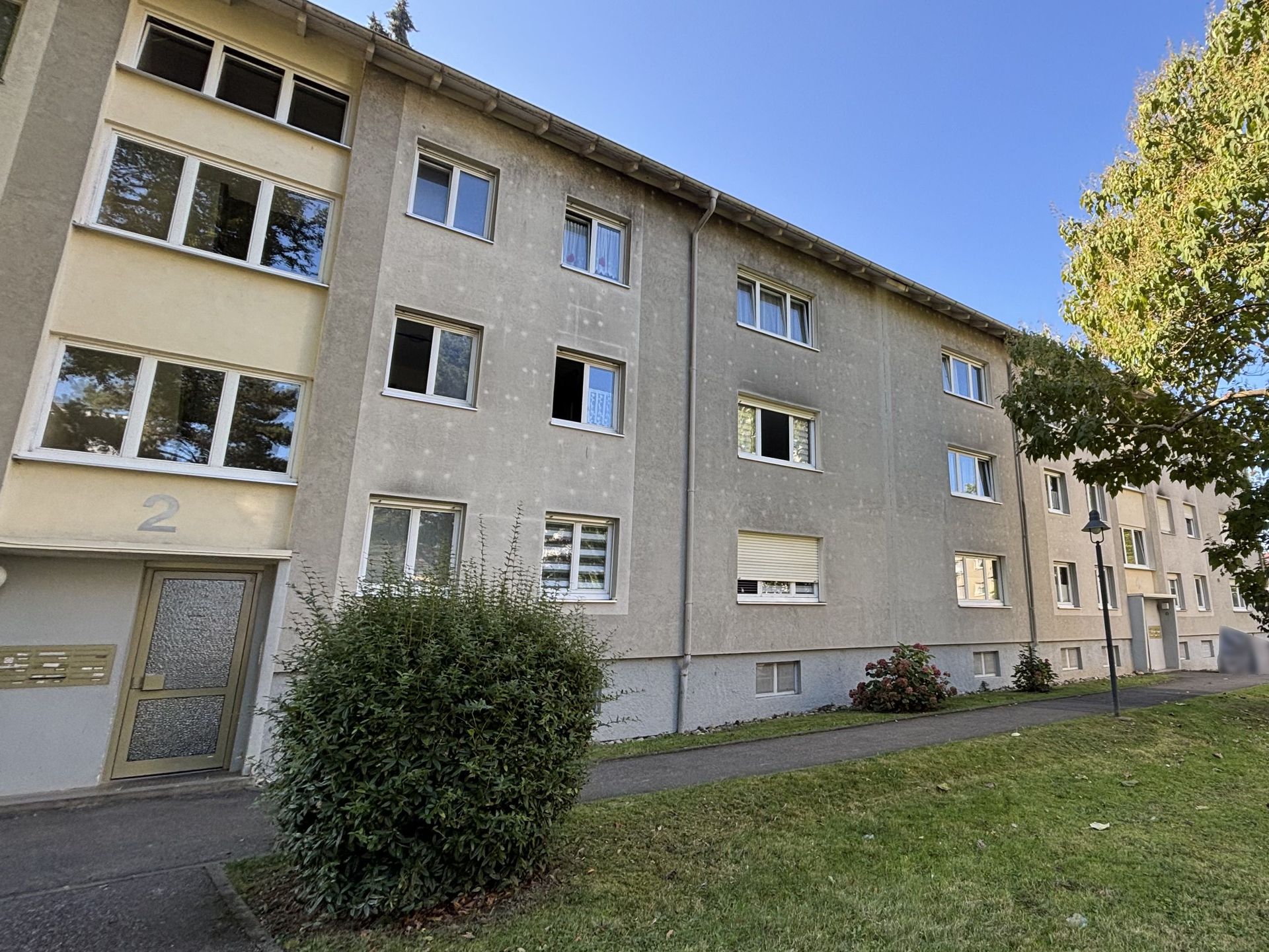 Das Bild zeigt ein Mehrfamilienhaus mit heller Fassade. Vor dem Haus befindet sich eine Grünfläche mit Sträuchern und Bäumen.