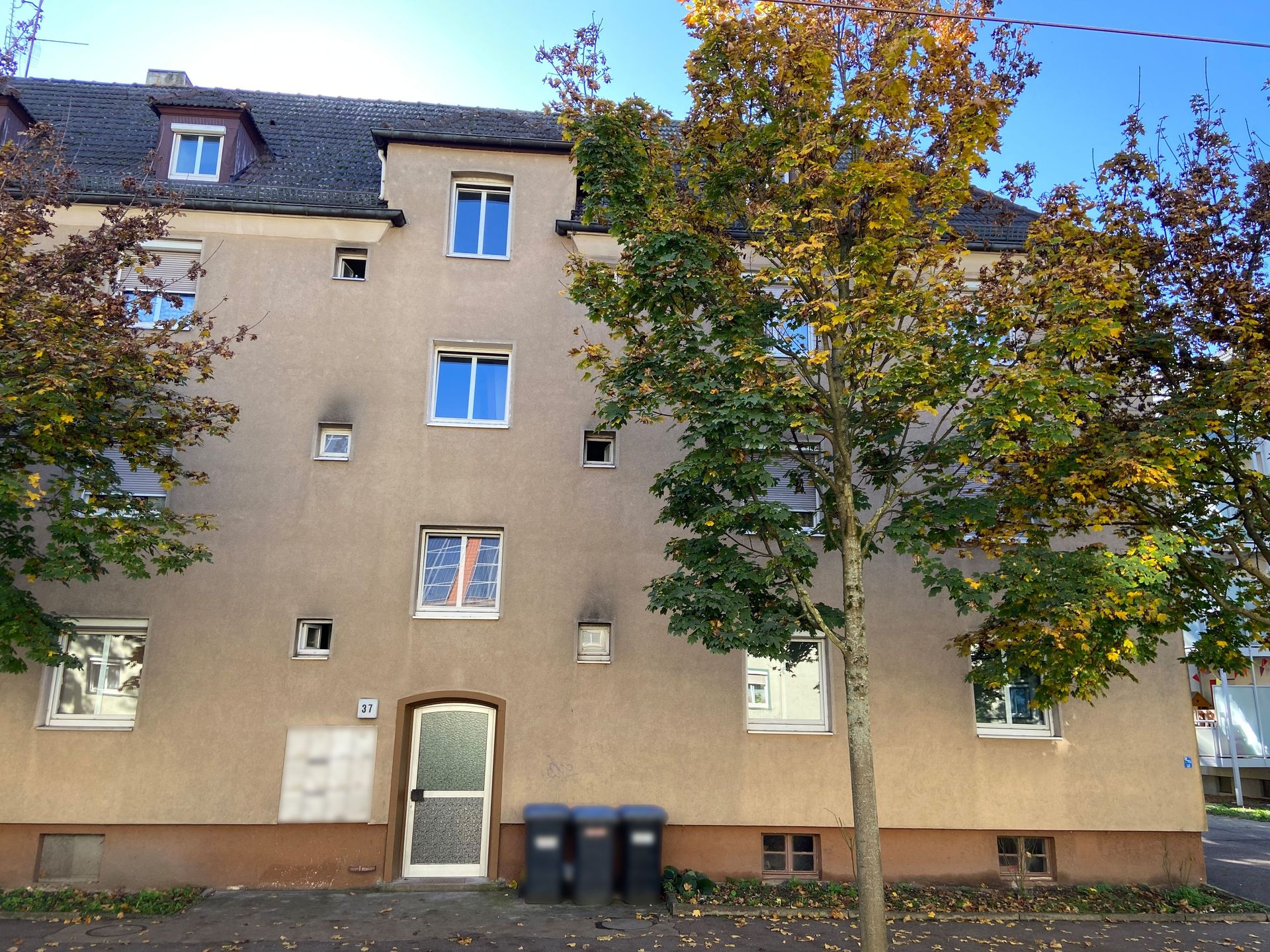 Das Bild zeigt ein Mehrfamilienhaus mit heller Fassade und dunklem Dach. Vor dem Haus stehen zwei Mülltonnen und ein Baum mit herbstlich gefärbten Blättern.
