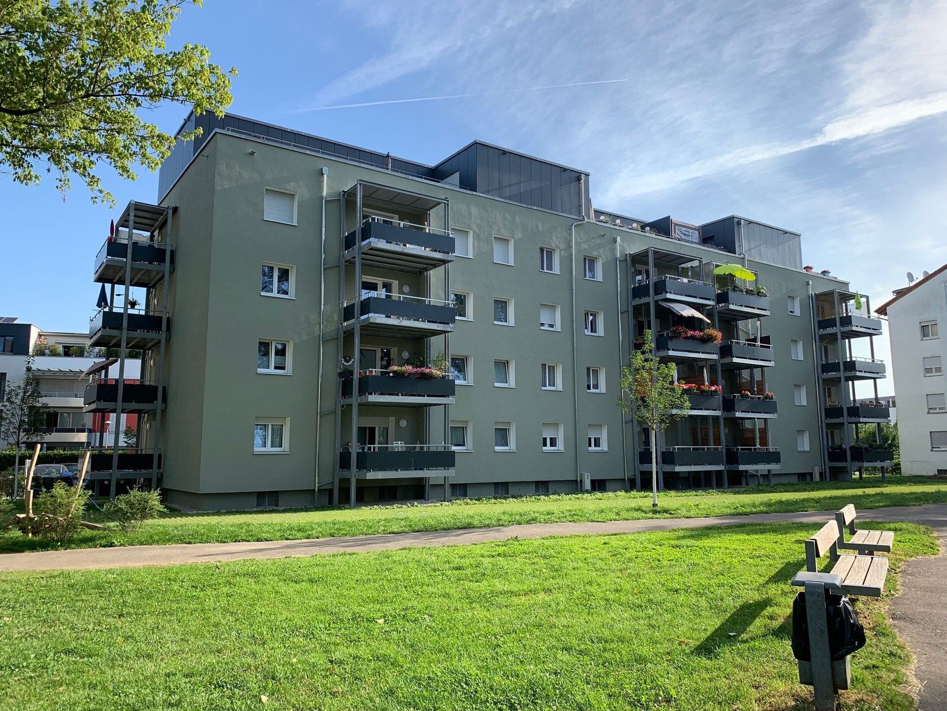 Das Bild zeigt ein Mehrfamilienhaus mit Balkonen. Vor dem Haus ist eine große Grünfläche mit zwei Sitzmöglichkeiten.