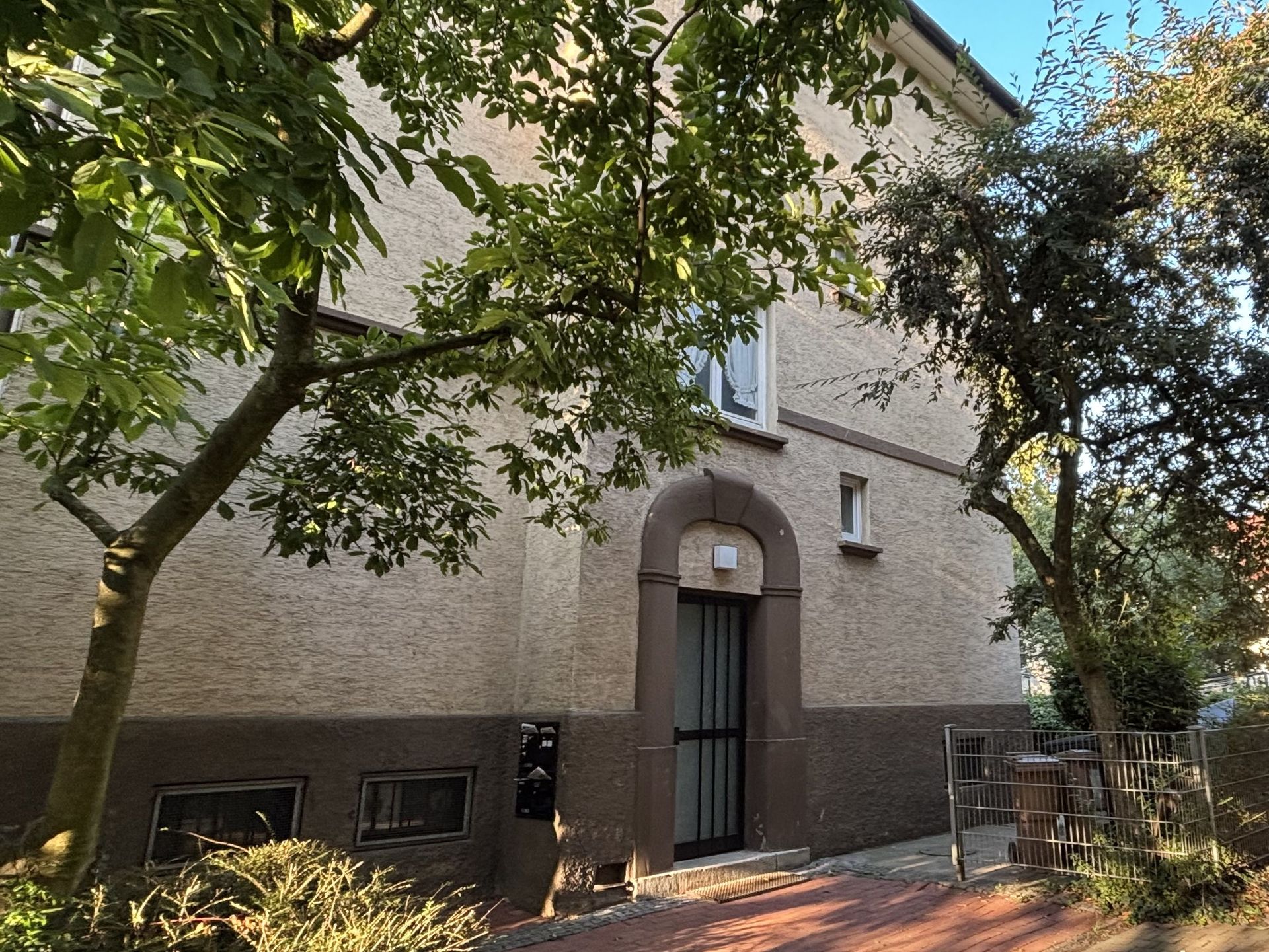 Das Bild zeigt ein Mehrfamilienhaus mit heller Fassade, das teilweise von Bäumen verdeckt wird.  Vor dem Haus befindet sich ein gepflasterter Weg.