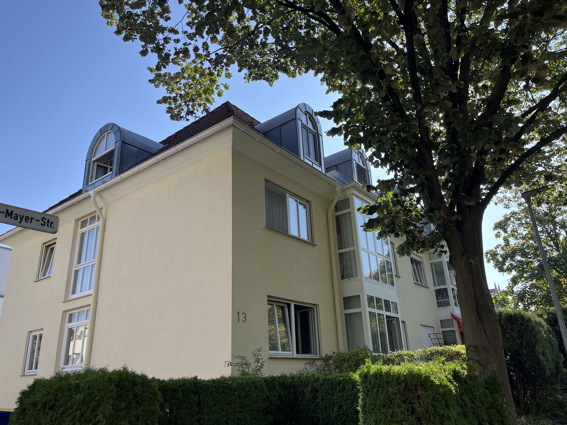 Das Bild zeigt ein helles Haus mit abgerundeten Dachgauben und großen Fenstern, die viel Licht hereinlassen.  Vor dem Haus stehen Bäume und eine Hecke.