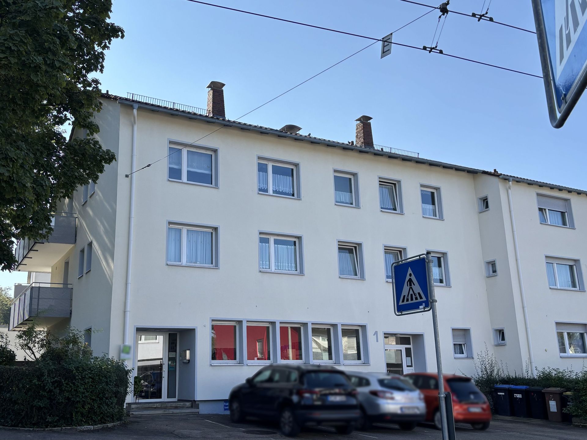 Das Bild zeigt ein Mehrfamilienhaus mit weißer Fassade und grauen Fensterrahmen.  Vor dem Haus befinden sich Parkplätze und ein Fußgängerüberweg.