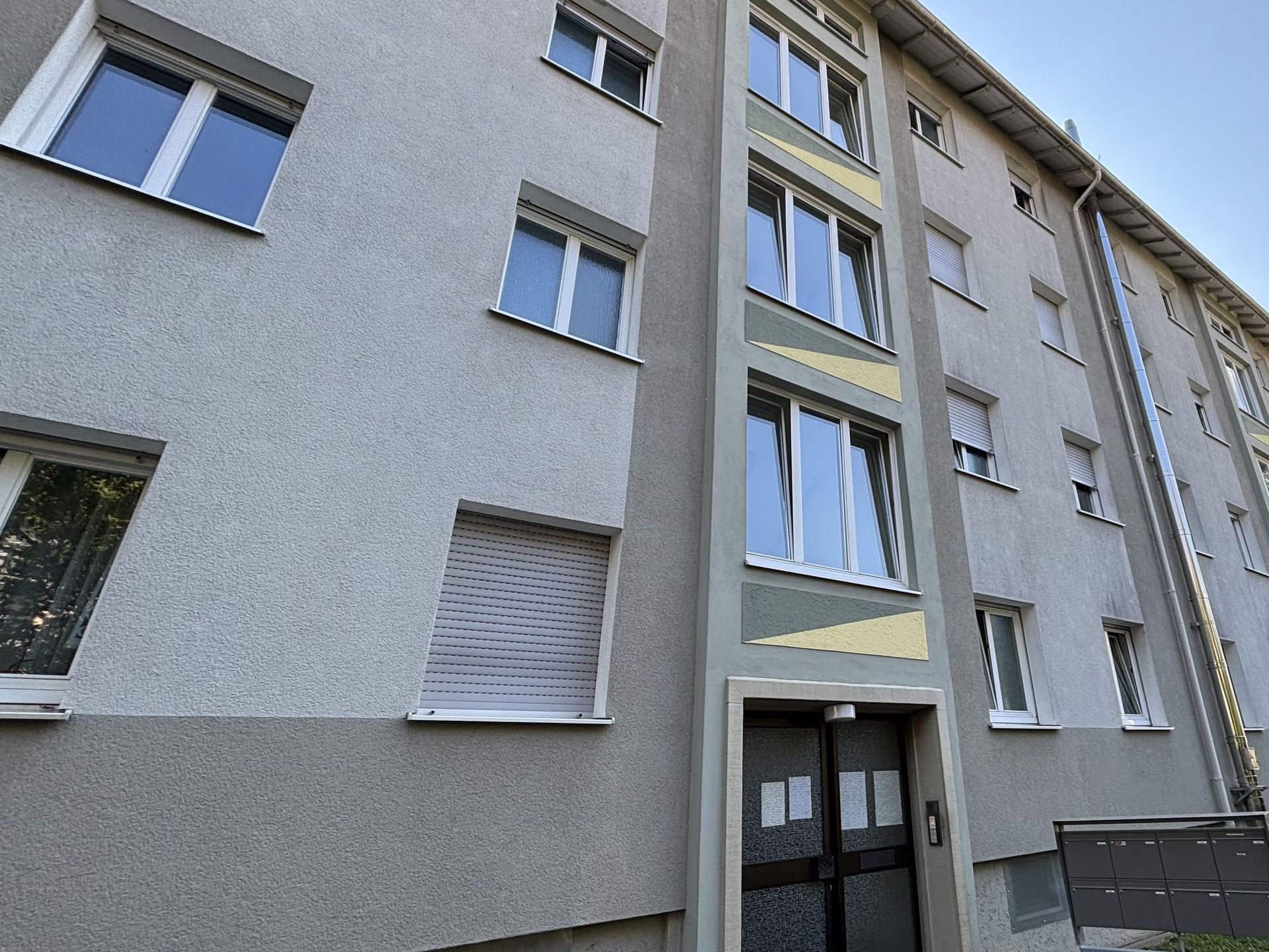 Das Bild zeigt ein Mehrfamilienhaus mit grauer Fassade, an der gelbe und grüne Farbakzente zwischen die Fenster gesetzt wurden. Das Gebäude hat mehrere Stockwerke.