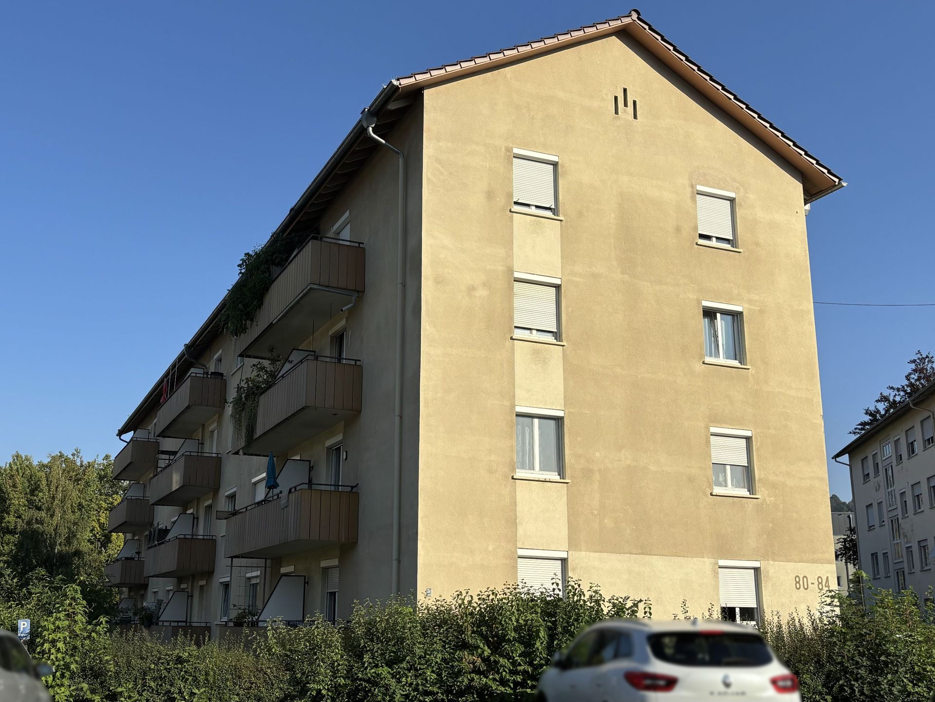 Das Bild zeigt ein Mehrfamilienhaus mit hellgelber Fassade und mehreren Balkonen. Vor dem Haus parkt ein Auto am Straßenrand und eine grüne Hecke begrenzt das Grundstück.