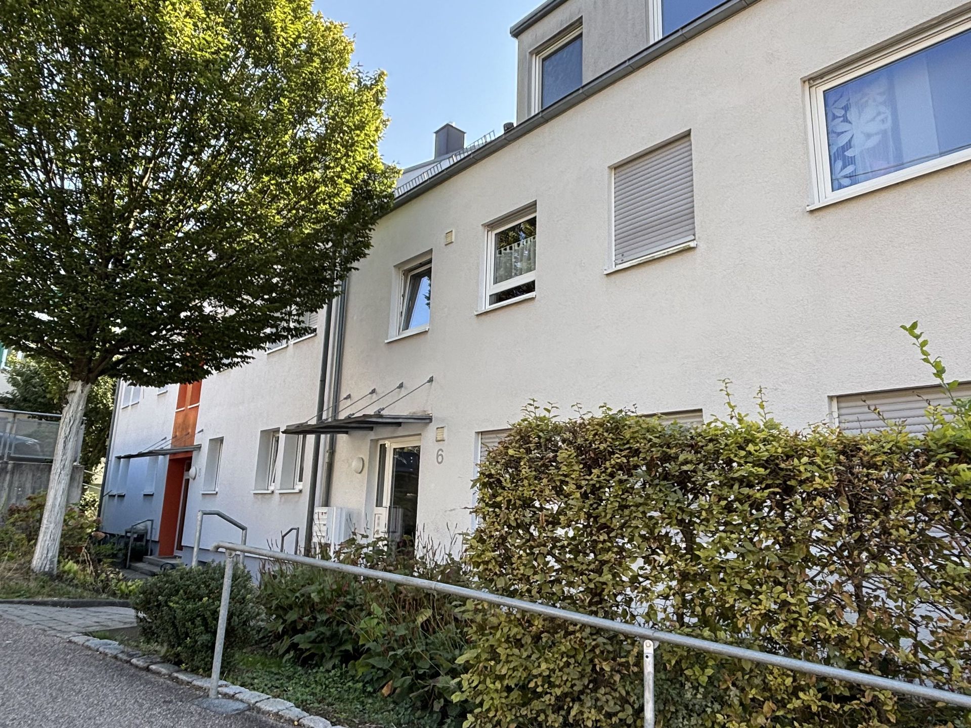 Das Bild zeigt ein Reihenhaus mit heller Fassade. Vor dem Haus befindet sich ein kleiner Vorgarten mit einem Baum und einer Hecke.