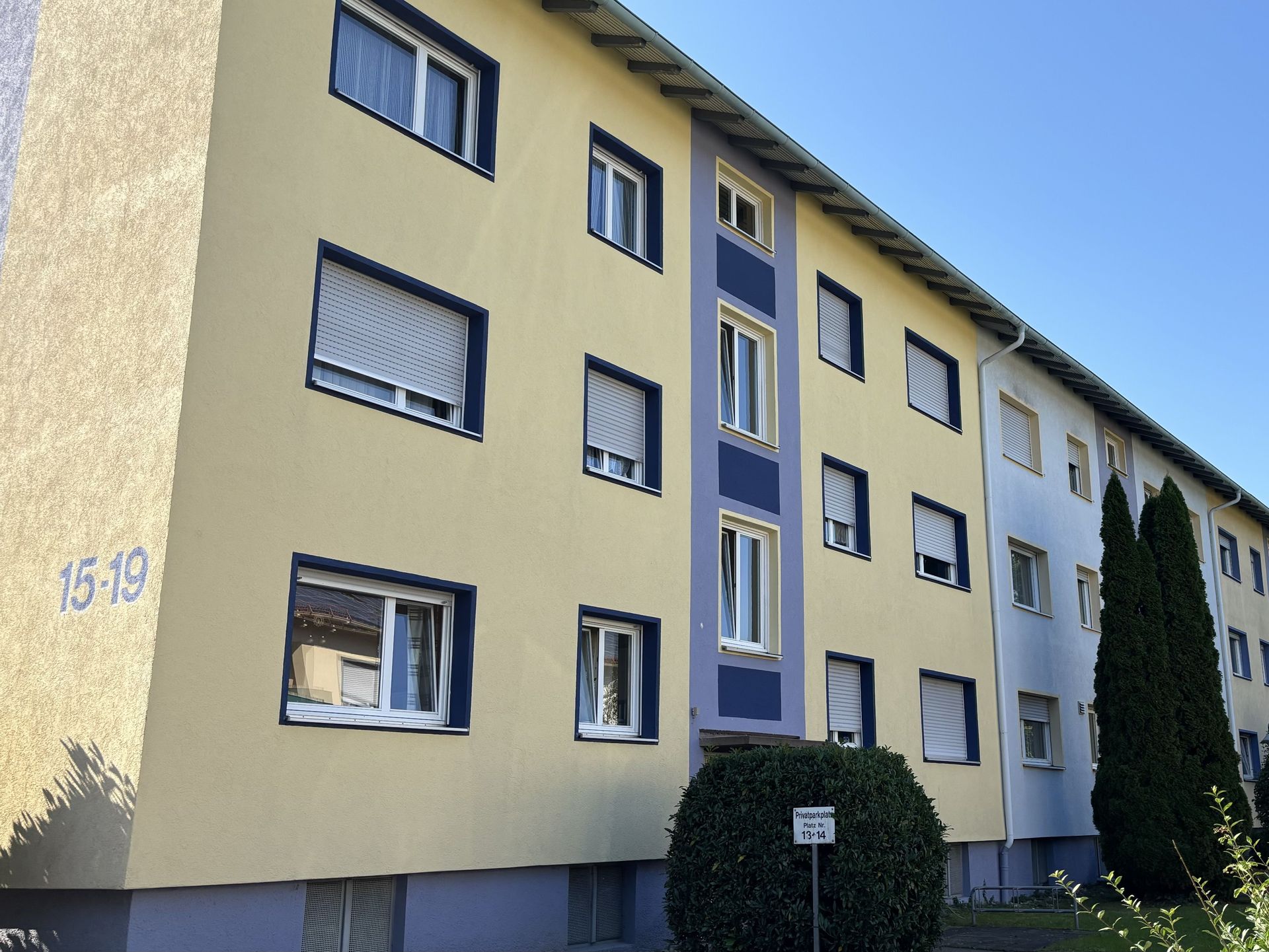 Das Bild zeigt ein Mehrfamilienhaus mit hellgelber Fassade und dunkelblauen Akzenten um die Fenster.  Vor dem Haus befinden sich Sträucher und ein kleiner Baum.