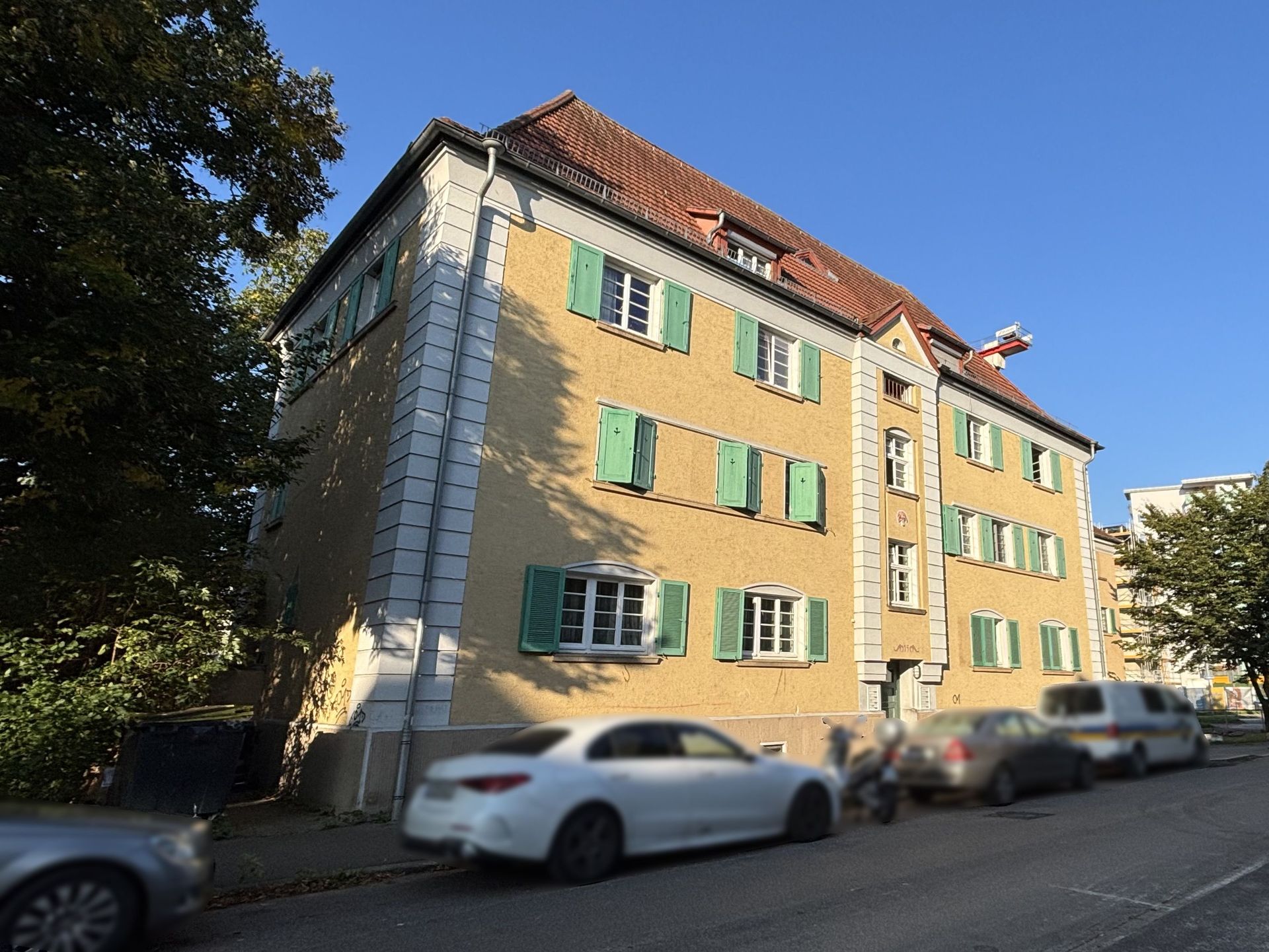 Das Bild zeigt ein Mehrfamilienhaus mit hellgelber Fassade und grünen Fensterläden. Vor dem Haus parken mehrere Autos am Straßenrand und ein Baum steht links im Bild.