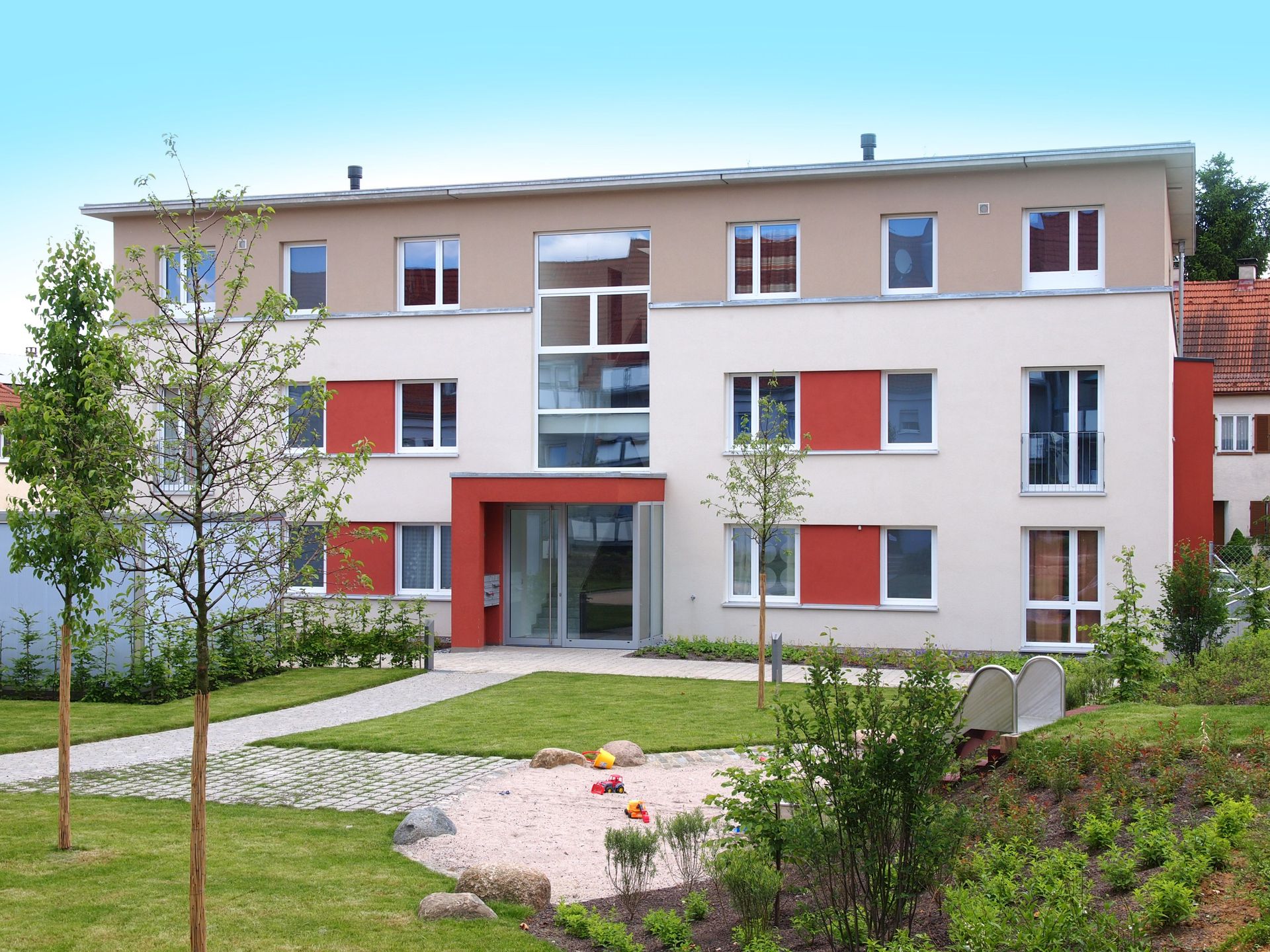 Das Bild zeigt ein modernes Mehrfamilienhaus mit heller Fassade, großen Fenstern und einigen roten Akzenten an den Fensterläden. Vor dem Haus befindet sich eine Grünfläche mit Spielplatz und Bäumen.
