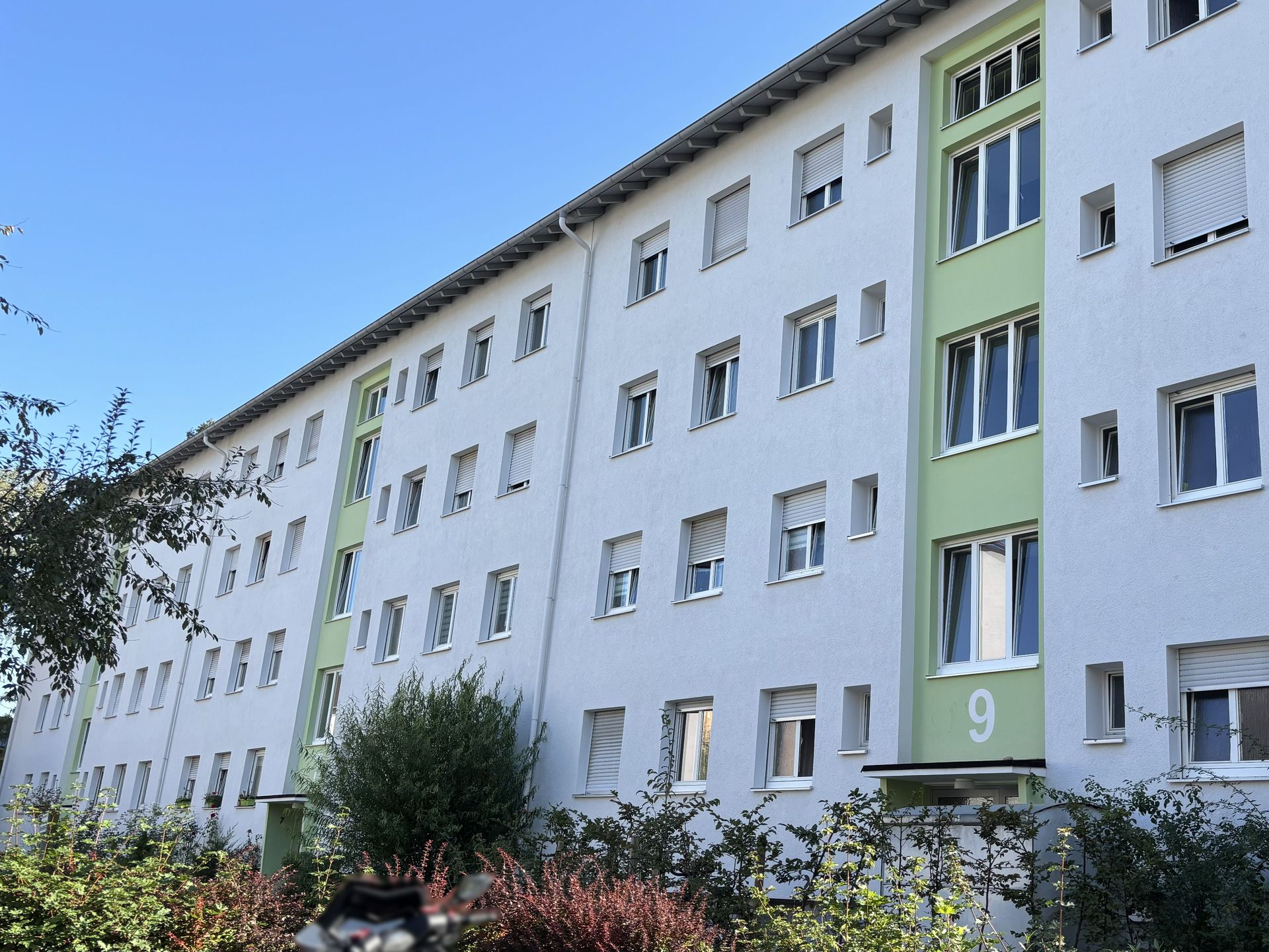 Das Bild zeigt ein Mehrfamilienhaus mit weißer Fassade und einigen hellgrünen Akzenten. Vor dem Gebäude befinden sich Sträucher und ein Baum.