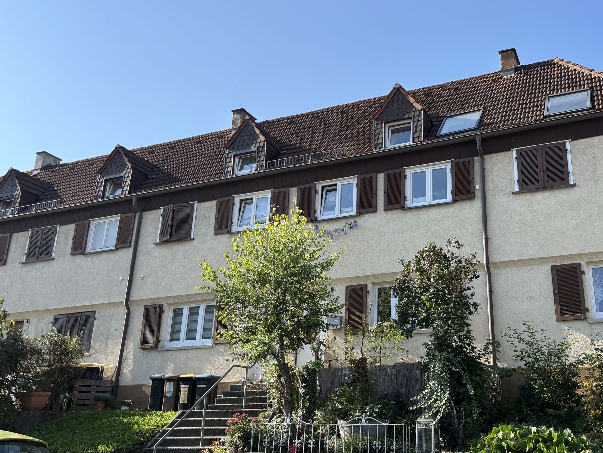 Das Bild zeigt eine Reihe von Häusern mit heller Fassade, braunem Dach und braunen Fensterläden. Vor den Häusern befinden sich kleine Vorgärten mit Sträuchern und Bäumen.