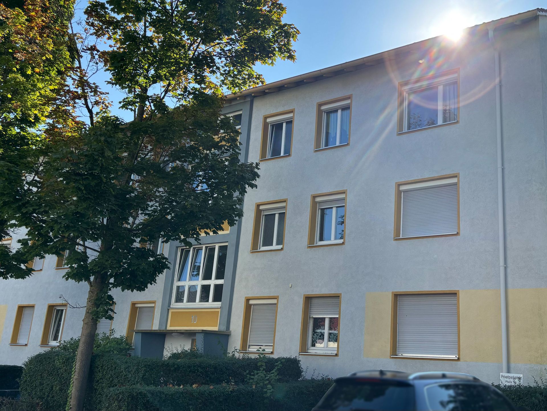 Das Bild zeigt ein Mehrfamilienhaus mit heller Fassade und farblich abgesetzten Fensterrahmen in Gelb.  Vor dem Haus stehen ein Baum und eine Hecke, und am Straßenrand ist ein Auto geparkt.