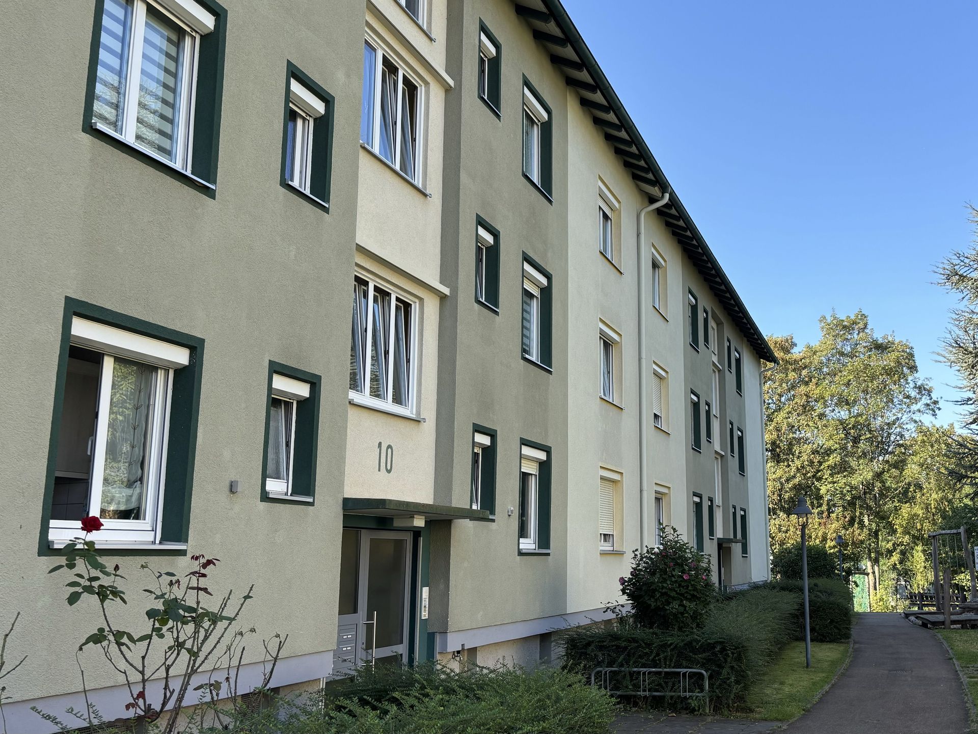 Das Bild zeigt ein Mehrfamilienhaus mit hellgrüner Fassade und dunkelgrünen Fensterläden. Neben dem Haus führt ein Weg entlang, der von einer Hecke und Bäumen gesäumt ist.