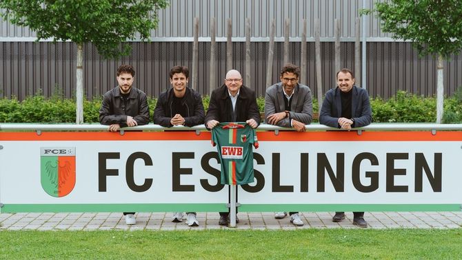 Das Bild zeigt fünf Personen, die hinter einer Bande auf einem Sportplatz stehen. Eine Person hält ein Trikot mit dem EWB-Logo drauf.