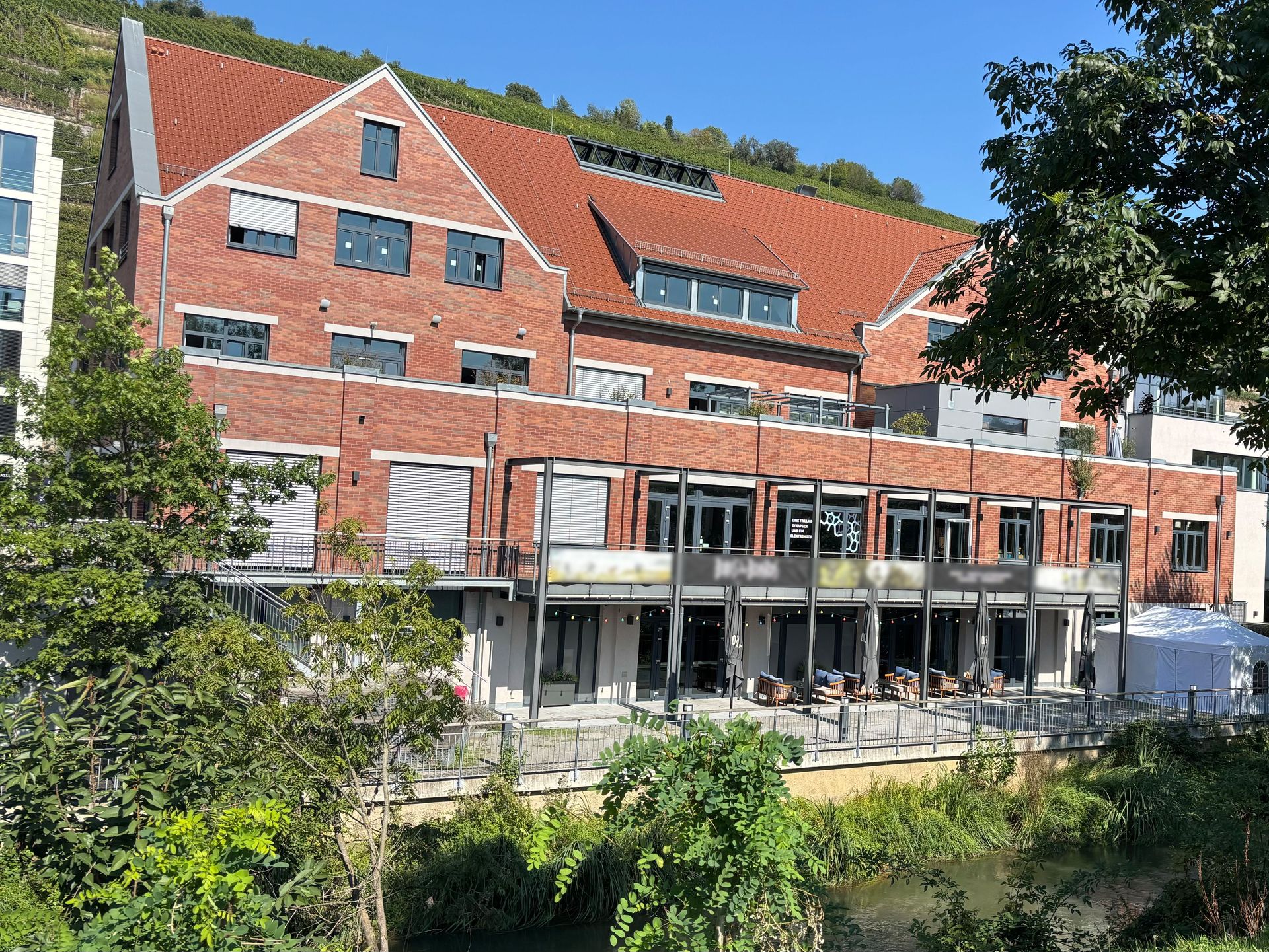 Das Bild zeigt ein modernes Gebäude mit roter Klinkerfassade und großen Fensterfronten im Erdgeschoss, die sich zu einer Terrasse hin öffnen. Das Gebäude liegt direkt an einem Fluss und ist von Bäumen und Sträuchern umgeben.