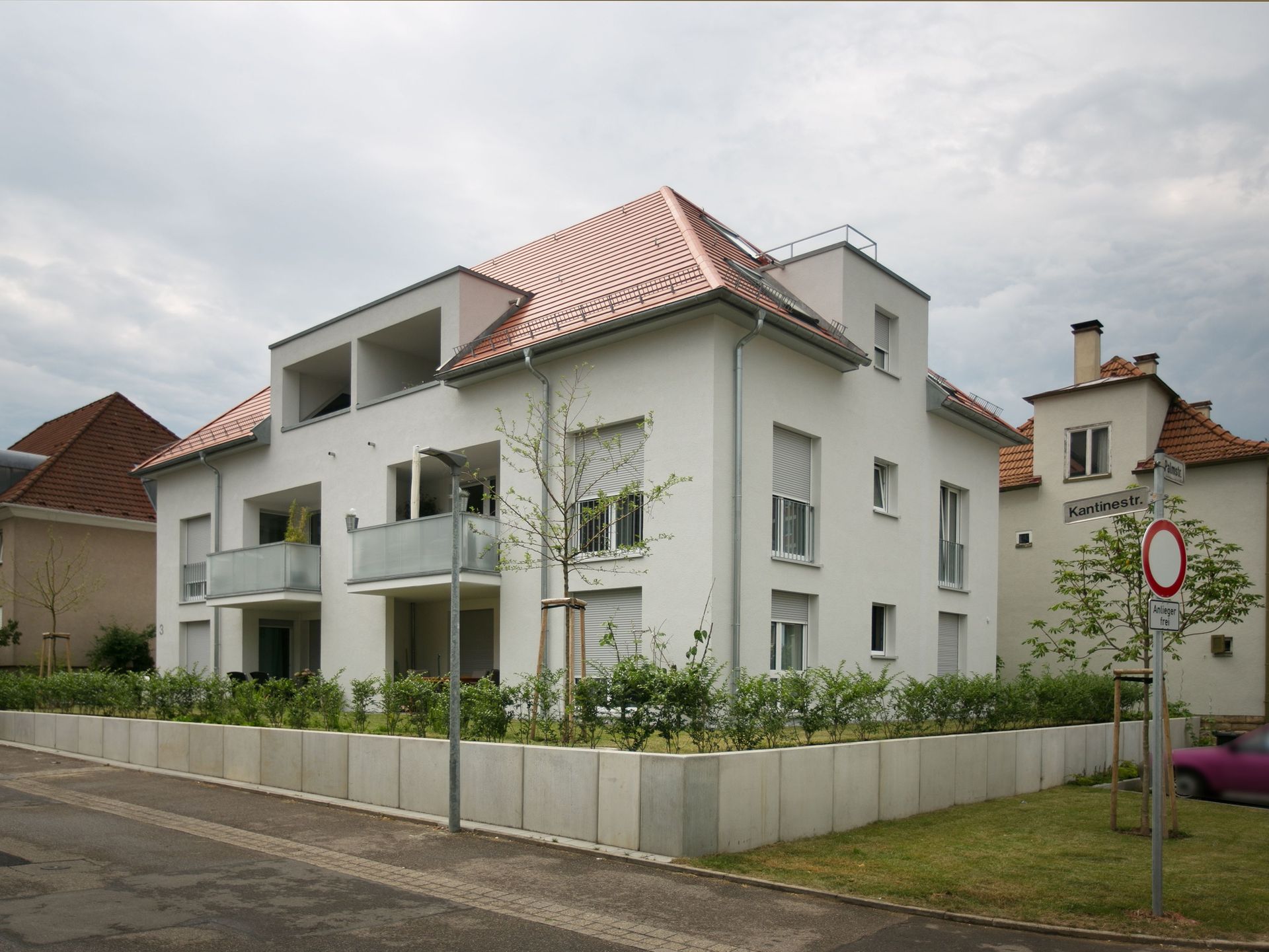 Das Bild zeigt ein modernes Mehrfamilienhaus mit weißer Fassade, großen Fenstern und einem roten Ziegeldach. Das Haus grenzt an eine ruhige Straße.