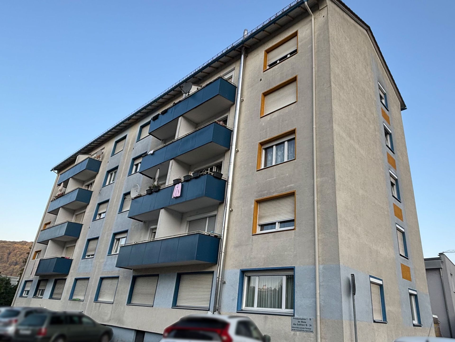 Das Bild zeigt ein Mehrfamilienhaus mit heller Fassade und blauen Balkonen, an dem einige Fenster mit orangefarbenen Akzenten ausgestattet sind. Vor dem Haus stehen mehrere Autos am Straßenrand.
