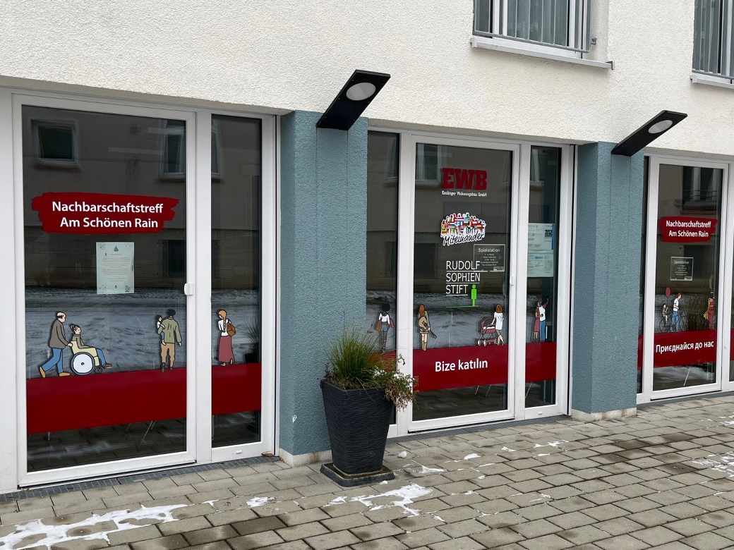 Das Bild zeigt einen Eingangsbereich eines Gebäudes. Die Fenster sind mit Gestaltungselementen beklebt und eine Pflanze steht dort. 