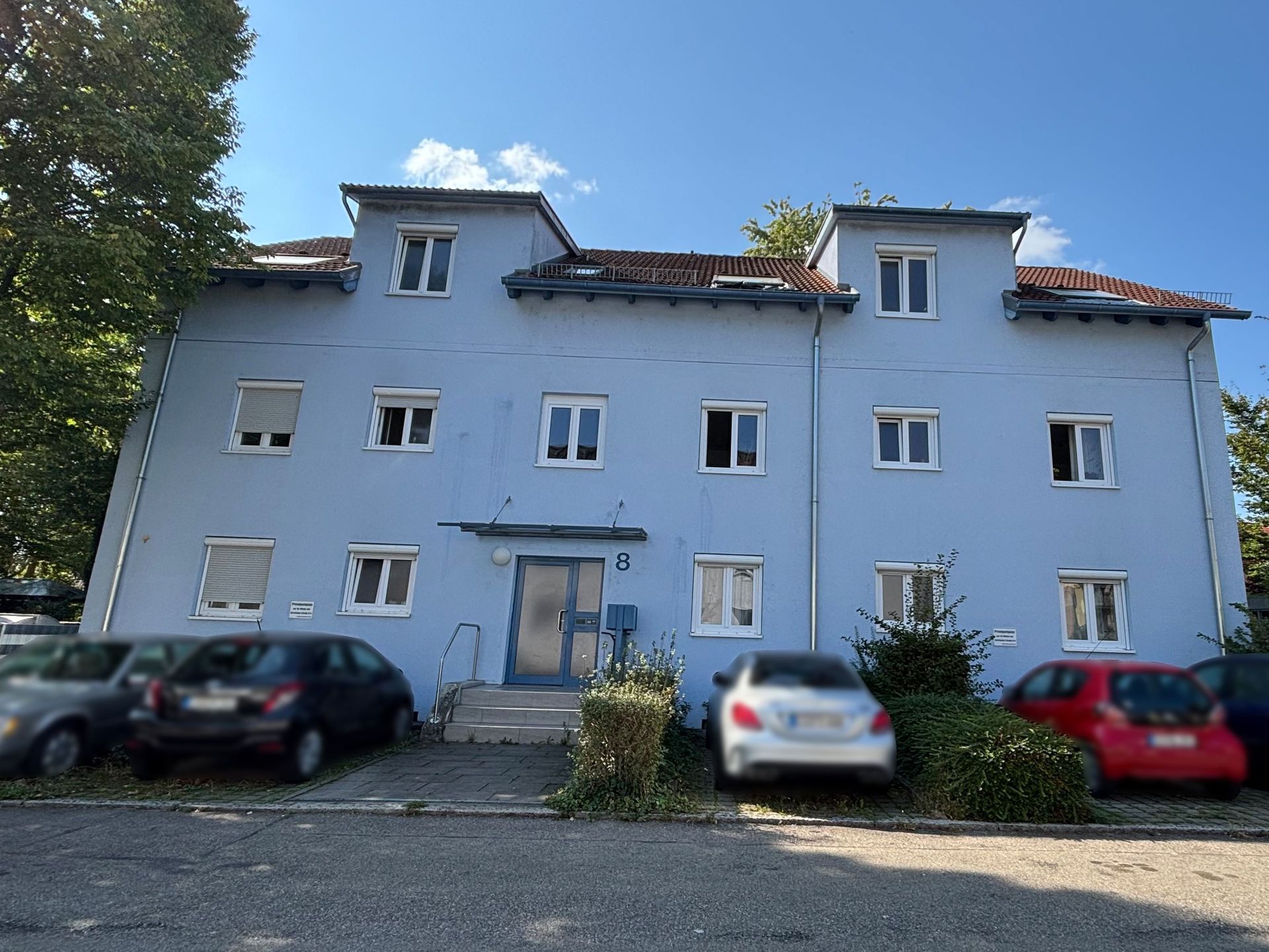 Das Bild zeigt ein hellblaues mehrstöckiges Haus. Vor dem Haus sind Sträucher und Autos auf Parkplätzen zu sehen.