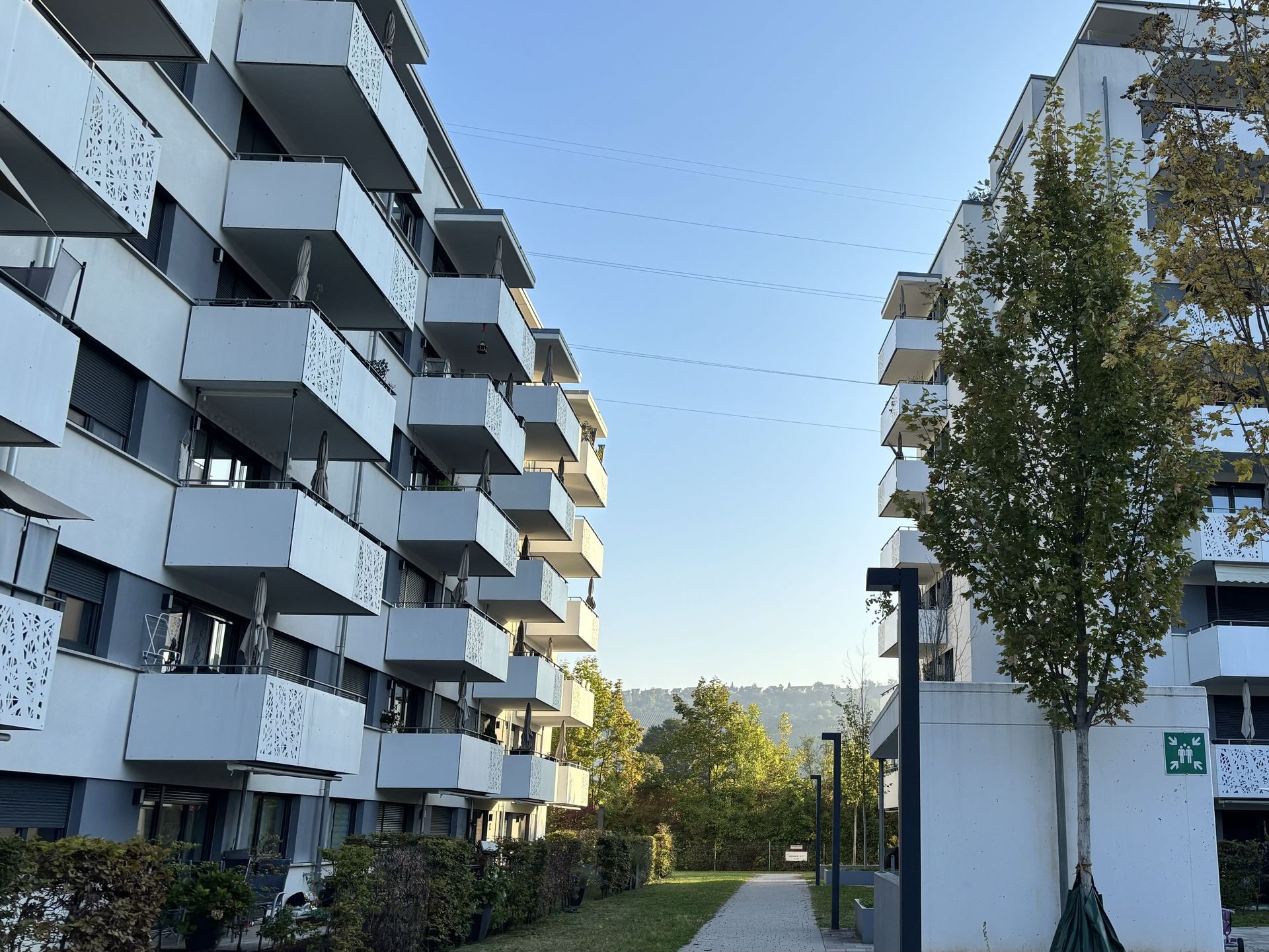 Das Bild zeigt zwei moderne Mehrfamilienhäuser mit Balkonen und Fassaden in Weißtönen. Zwischen den Gebäuden führt ein Weg durch eine begrünte Fläche mit Bäumen und Sträuchern.