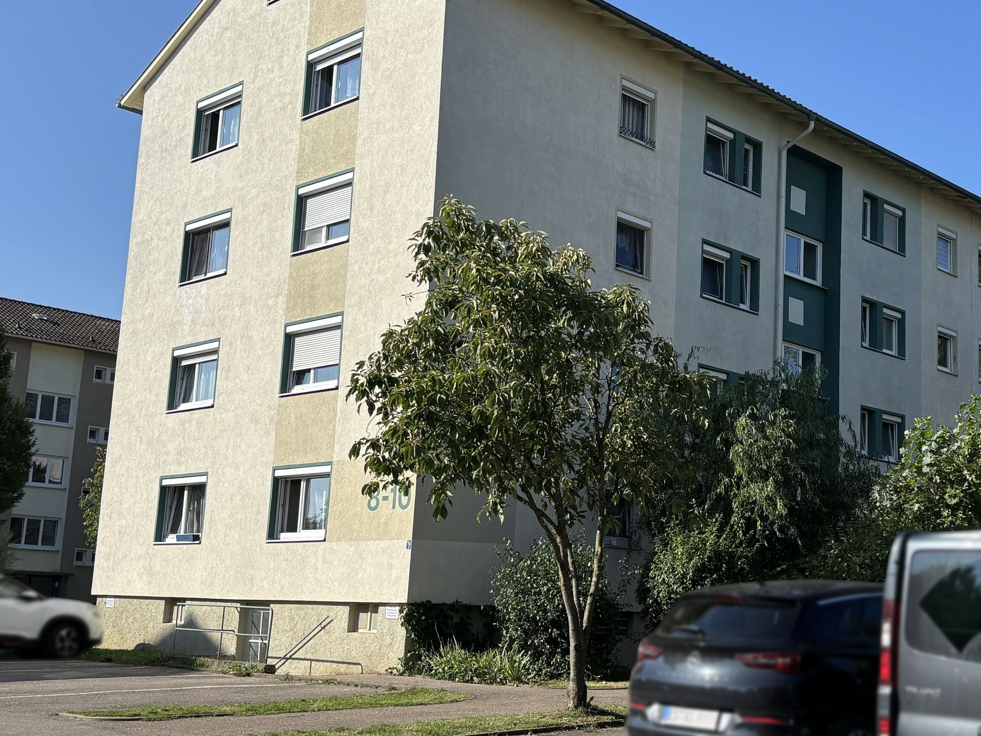Das Bild zeigt ein Mehrfamilienhaus mit heller Fassade. Vor dem Haus befinden sich Parkplätze, ein Baum und ein parkendes Auto.