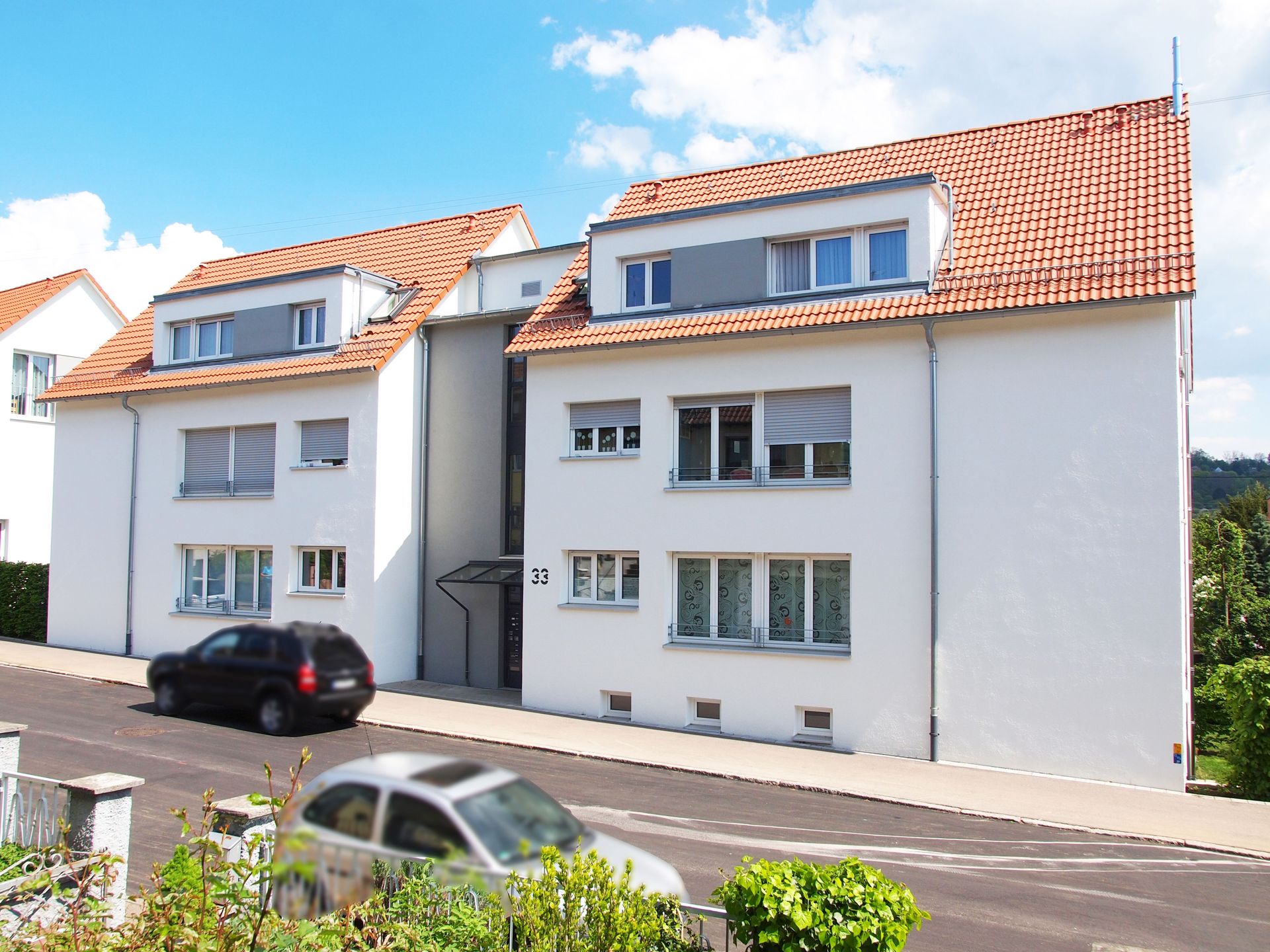 Das Bild zeigt zwei moderne Mehrfamilienhäuser mit weißer Fassade, roten Ziegeldächern und großen Fenstern. Die Häuser stehen an einer ruhigen Straße mit geparkten Autos und etwas Grün am Straßenrand.