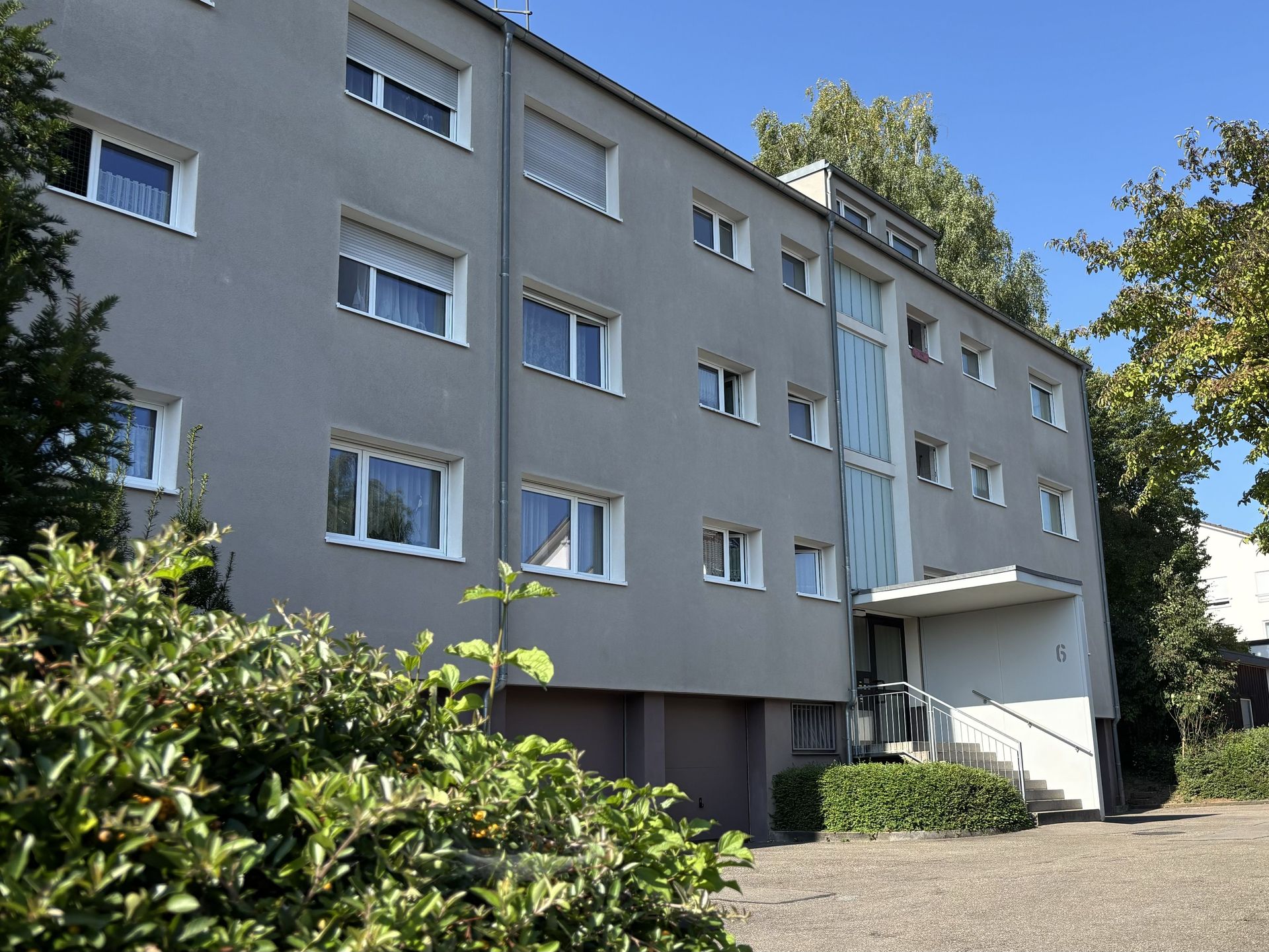 Das Bild zeigt ein Mehrfamilienhaus mit grauer Fassade. Vor dem Haus befinden sich eine kleine Treppe und einige Sträucher.
