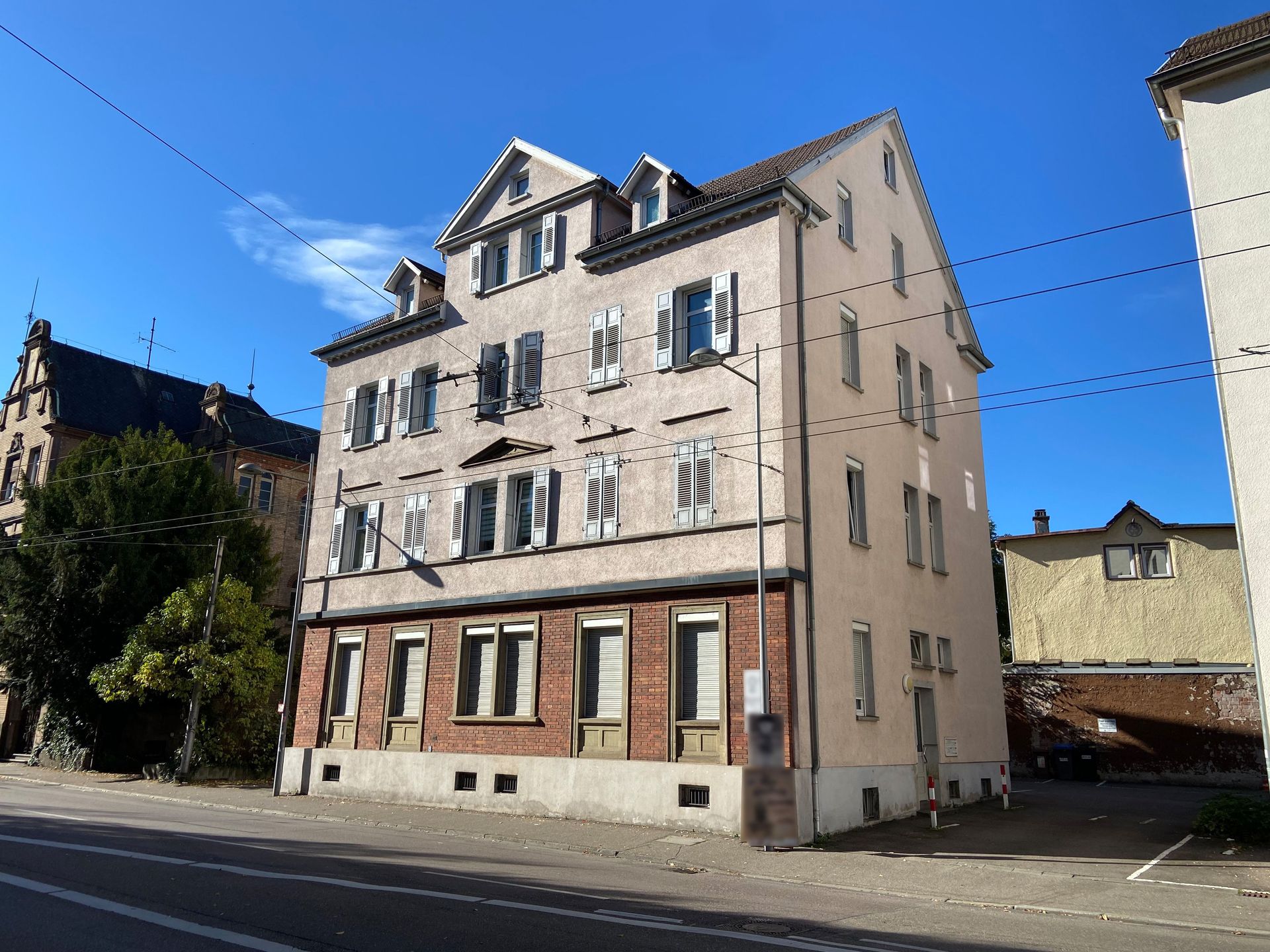 Das Bild zeigt ein Mehrfamilienhaus mit heller Fassade und weißen Fensterläden. Das Gebäude steht an einer Straße mit Oberleitungen für Straßenbahnen, und hat mehrere Stockwerke mit zahlreichen Fenstern.