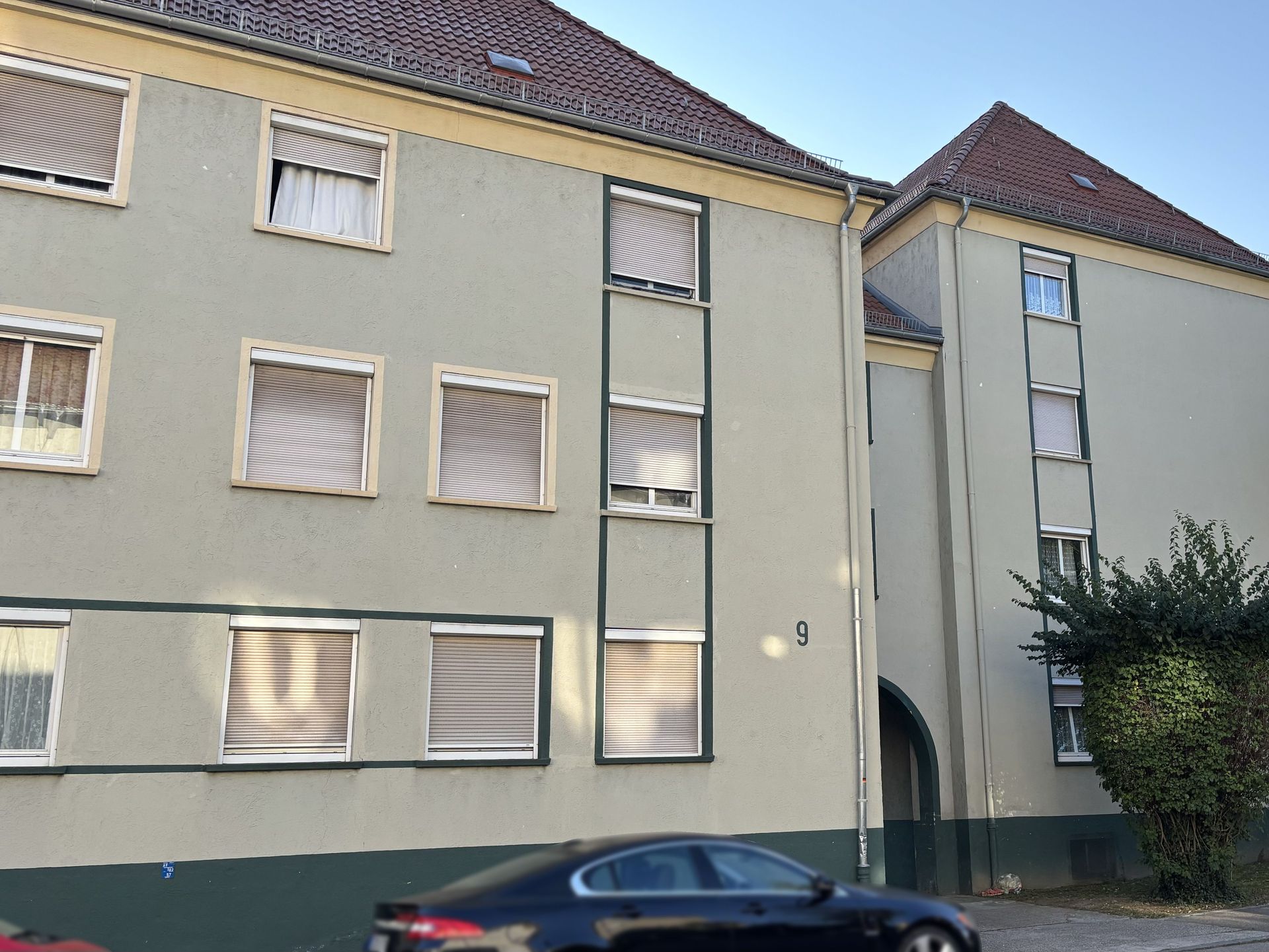 Das Bild zeigt ein Mehrfamilienhaus mit hellgrüner Fassade und abgesetzten, dunklen Fensterrahmen.  Vor dem Haus parkt ein Auto und rechts im Bild steht ein kleiner Baum.