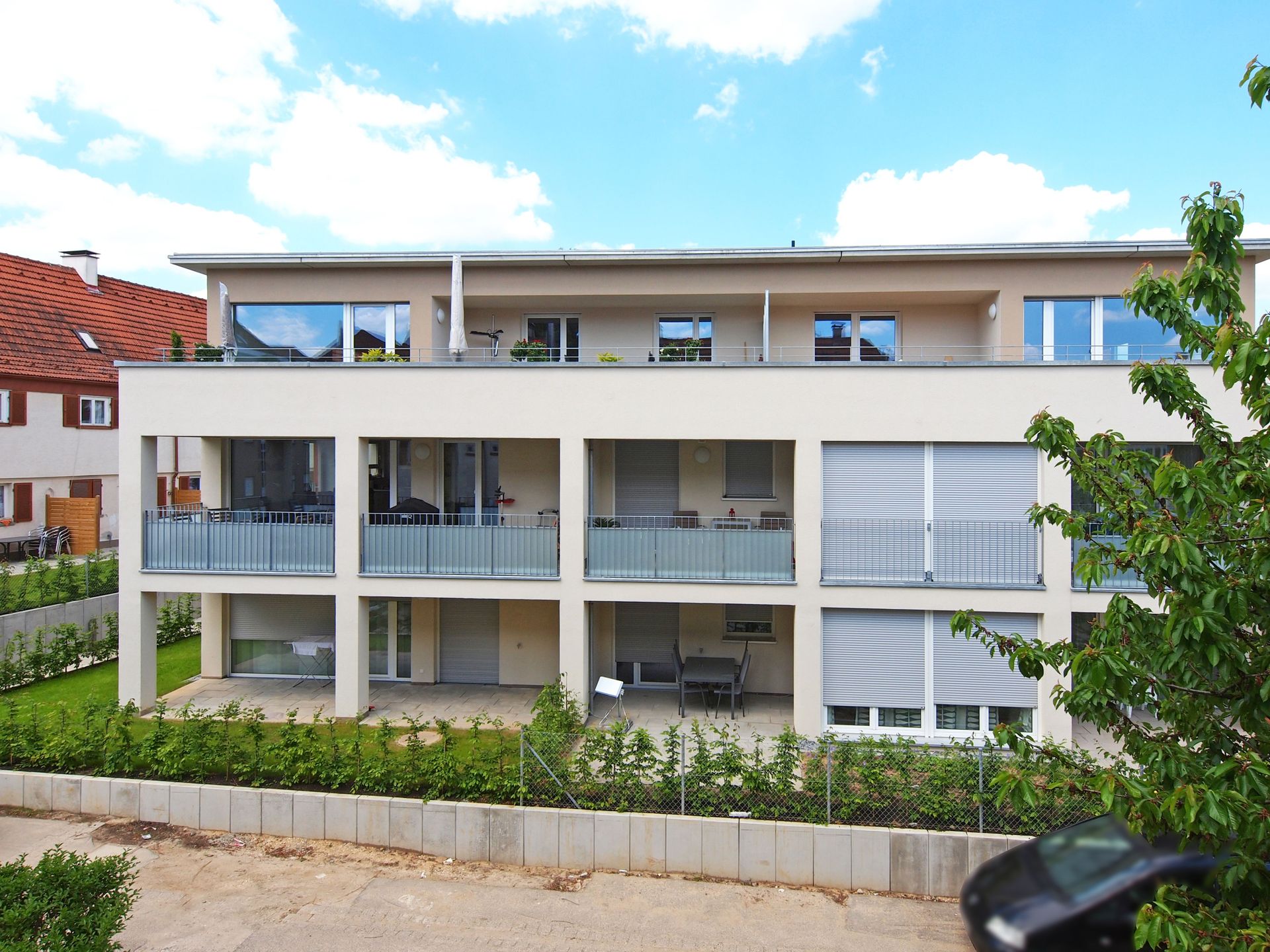 Das Bild zeigt ein modernes Mehrfamilienhaus mit heller Fassade, großen Fenstern und großzügigen Balkonen. Vor dem Haus befindet sich ein Garten mit einer niedrigen Hecke und im Hintergrund sind weitere Wohngebäude zu sehen.
