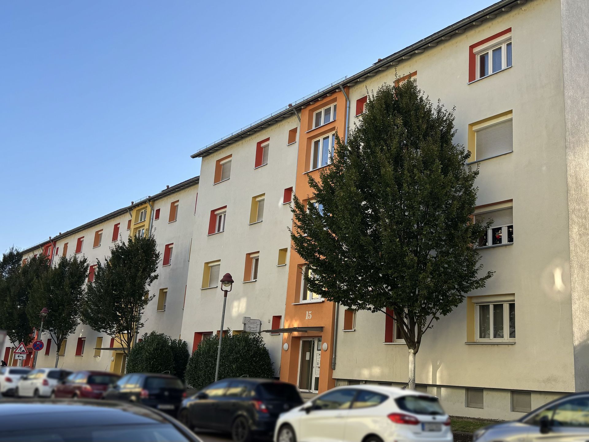 Das Bild zeigt ein Mehrfamilienhaus mit heller Fassade und farbigen Akzenten an den Fenstern und Balkonen. Vor dem Haus stehen mehrere Bäume und am Straßenrand sind Autos geparkt.