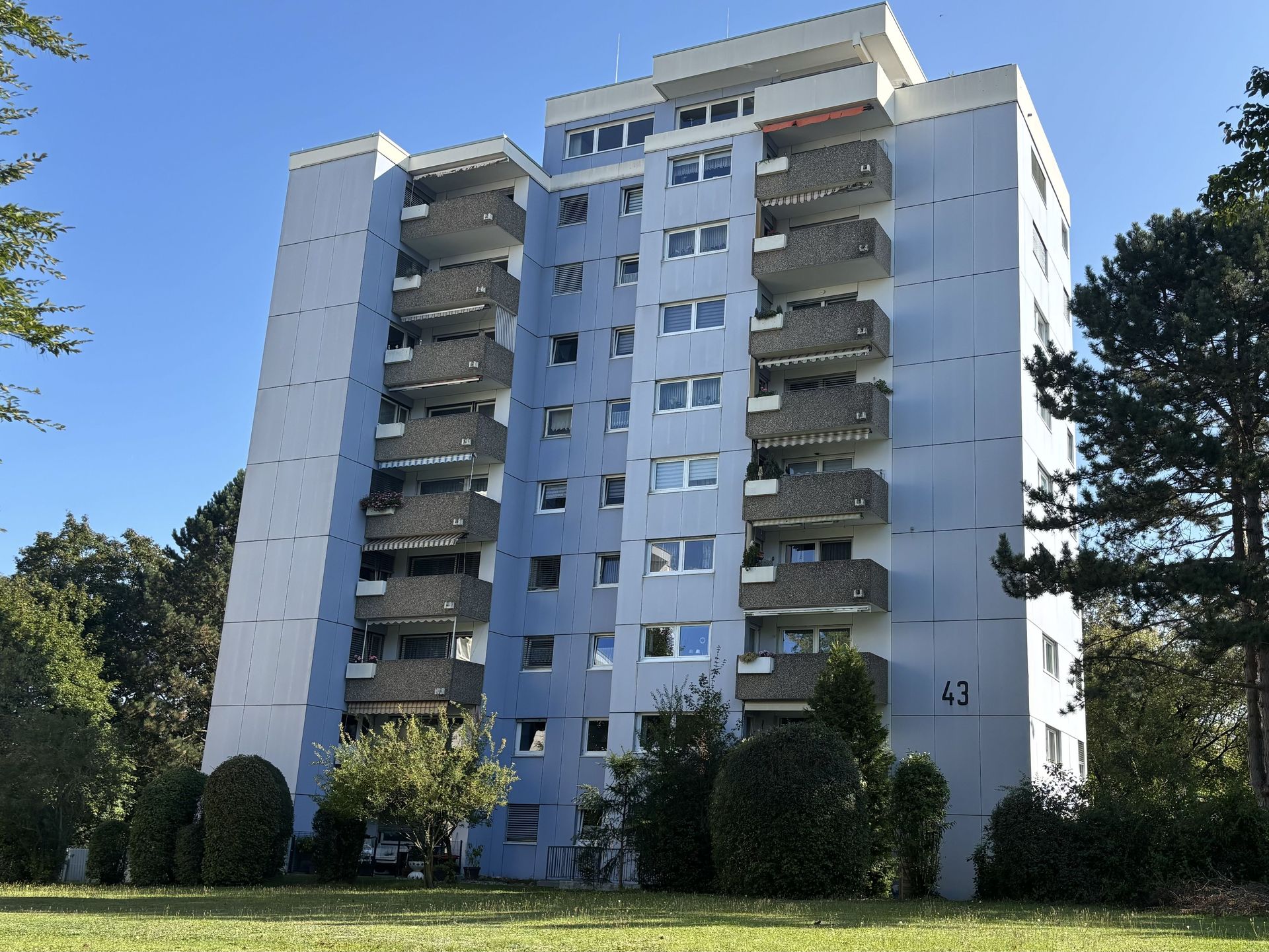 Das Bild zeigt ein hellblaues Hochhaus mit vielen Balkonen und Fensterreihen, das vor einem Hintergrund aus Bäumen steht. Vor dem Gebäude erstreckt sich eine gepflegte Rasenfläche, die von Büschen und Bäumen begrenzt wird.