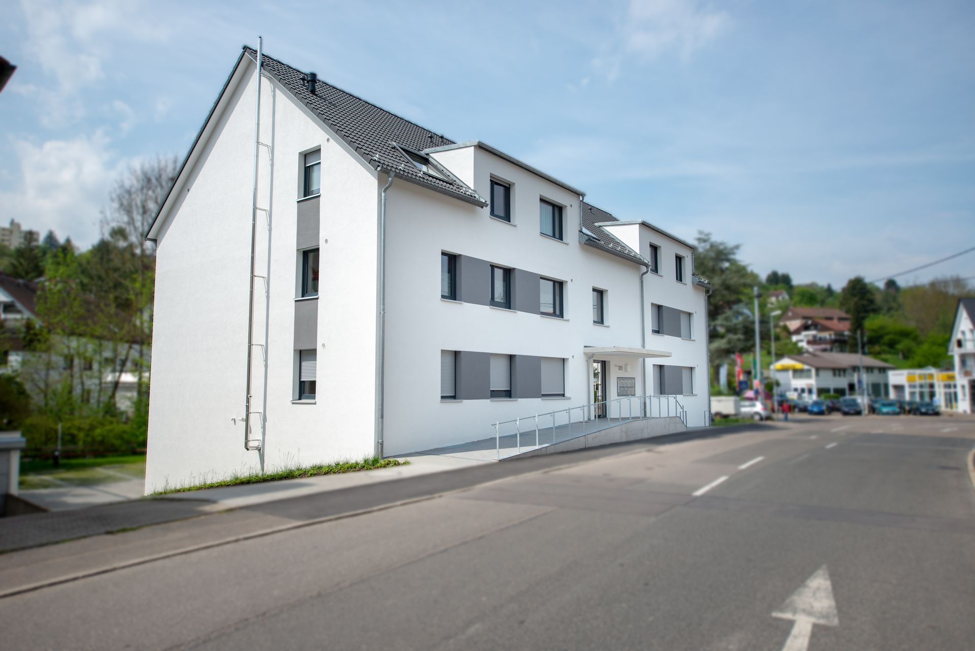 Das Bild zeigt ein modernes, mehrstöckiges Wohnhaus mit weißer Fassade und grauen Fensterläden.  Vor dem Haus befindet sich eine Rampe für einen barrierefreien Zugang.
